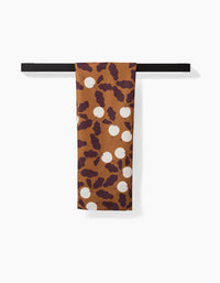 A rectangular brown Radishes kitchen tea towel by Lizzie Clark, featuring a maroon abstract pattern and white circles, hangs neatly over a horizontal black bar against a white background.