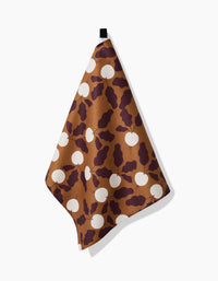 A brown, eco-friendly Radishes tea towel by Lizzie Clark features dark maroon leaf patterns and white circles, hanging from a black loop against a white background.