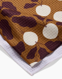 A close-up of the Lizzie Clark Radishes tea towel showcases brown and white tones with large circles and abstract dark purple shapes, plus a glimpse of its soft, absorbent reverse side.