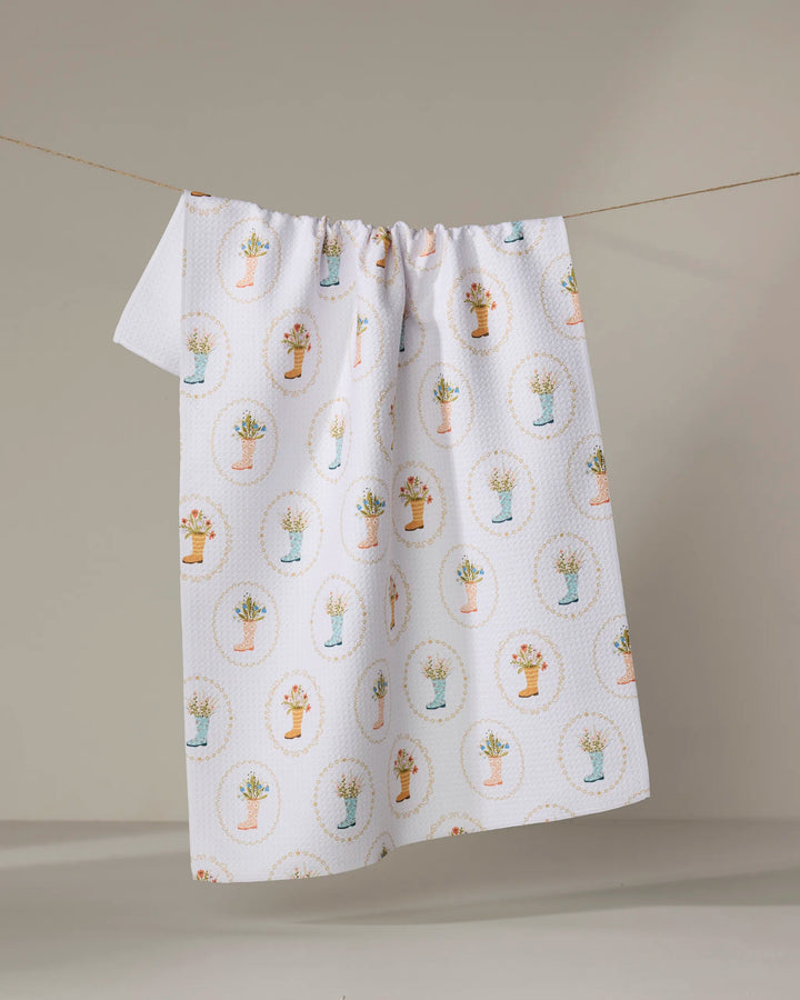 The Sara Mari "Rainboots in Bloom" tea towel, crafted from recycled materials and featuring colorful flower pots in oval frames, hangs on a clothesline against a neutral background.