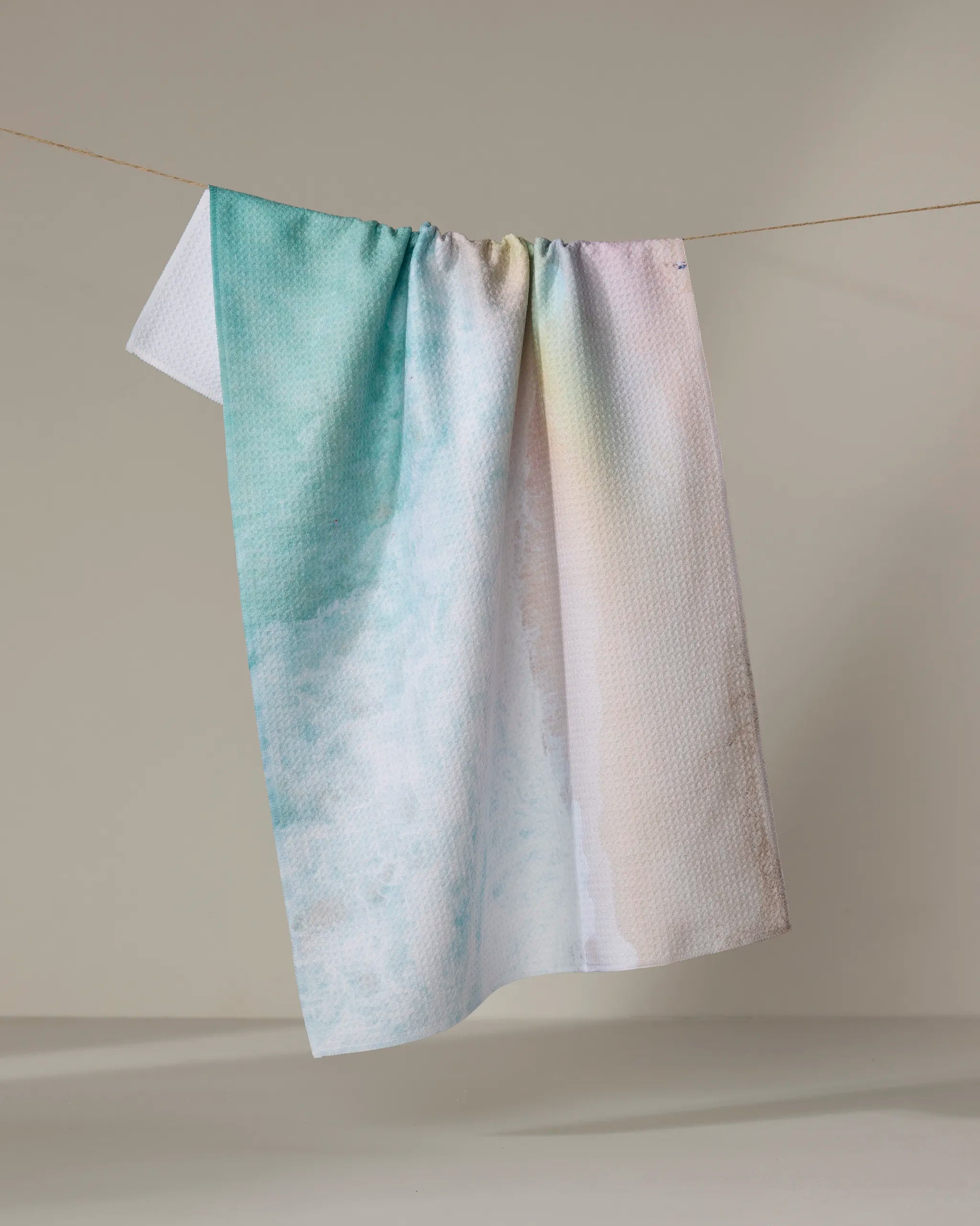 The Rainbow Wave by Kait Hanson, an absorbent dish towel featuring pastel hues and a soft watercolor gradient, hangs on a clothesline against a simple neutral backdrop.