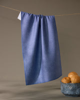 The Geometry Rainfield tea towel in textured blue hangs from a string against a plain background, with a ceramic bowl of three bread rolls placed below on the right.