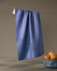 The Geometry Rainfield tea towel in textured blue hangs from a string against a plain background, with a ceramic bowl of three bread rolls placed below on the right.
