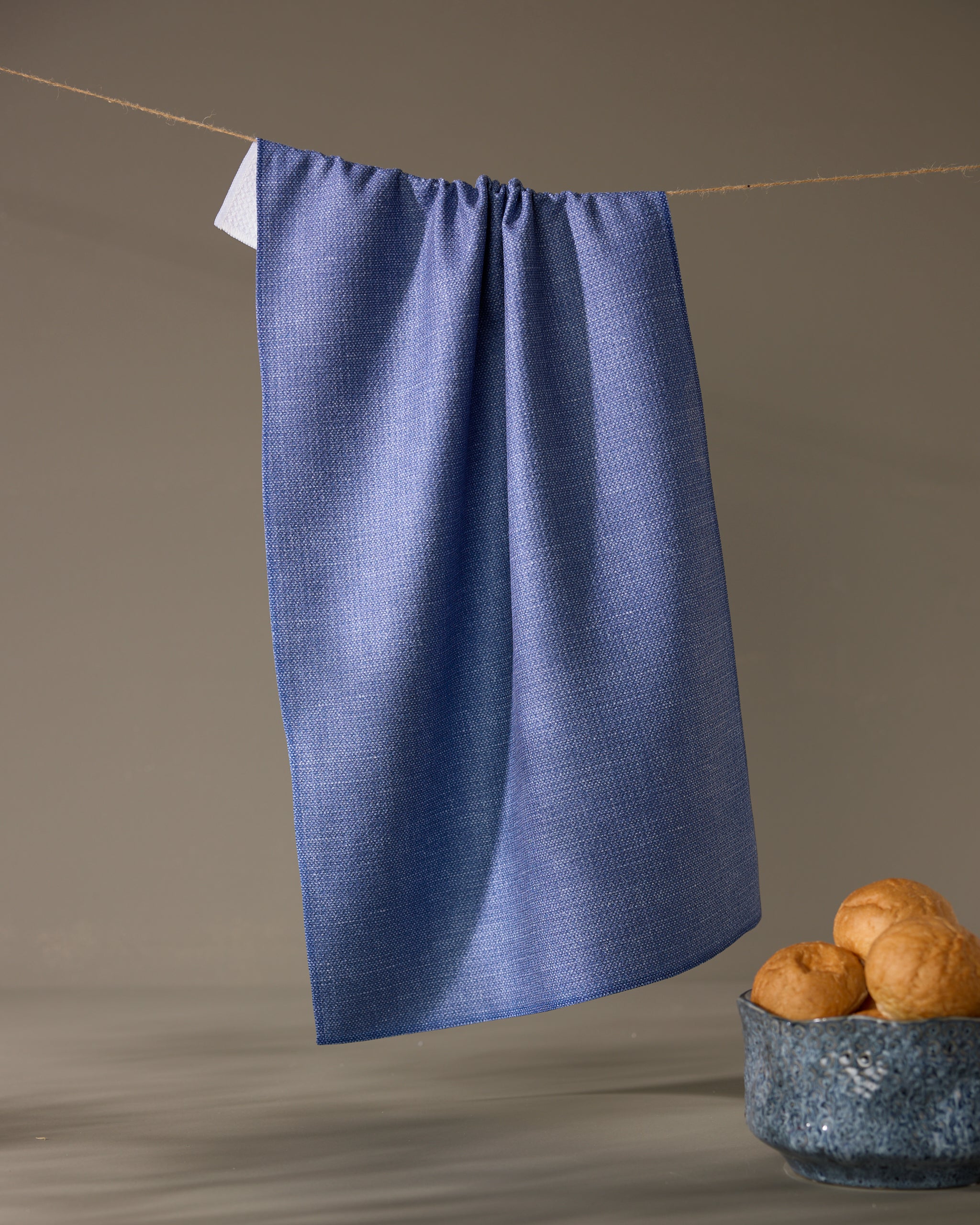 The Geometry Rainfield tea towel in textured blue hangs from a string against a plain background, with a ceramic bowl of three bread rolls placed below on the right.