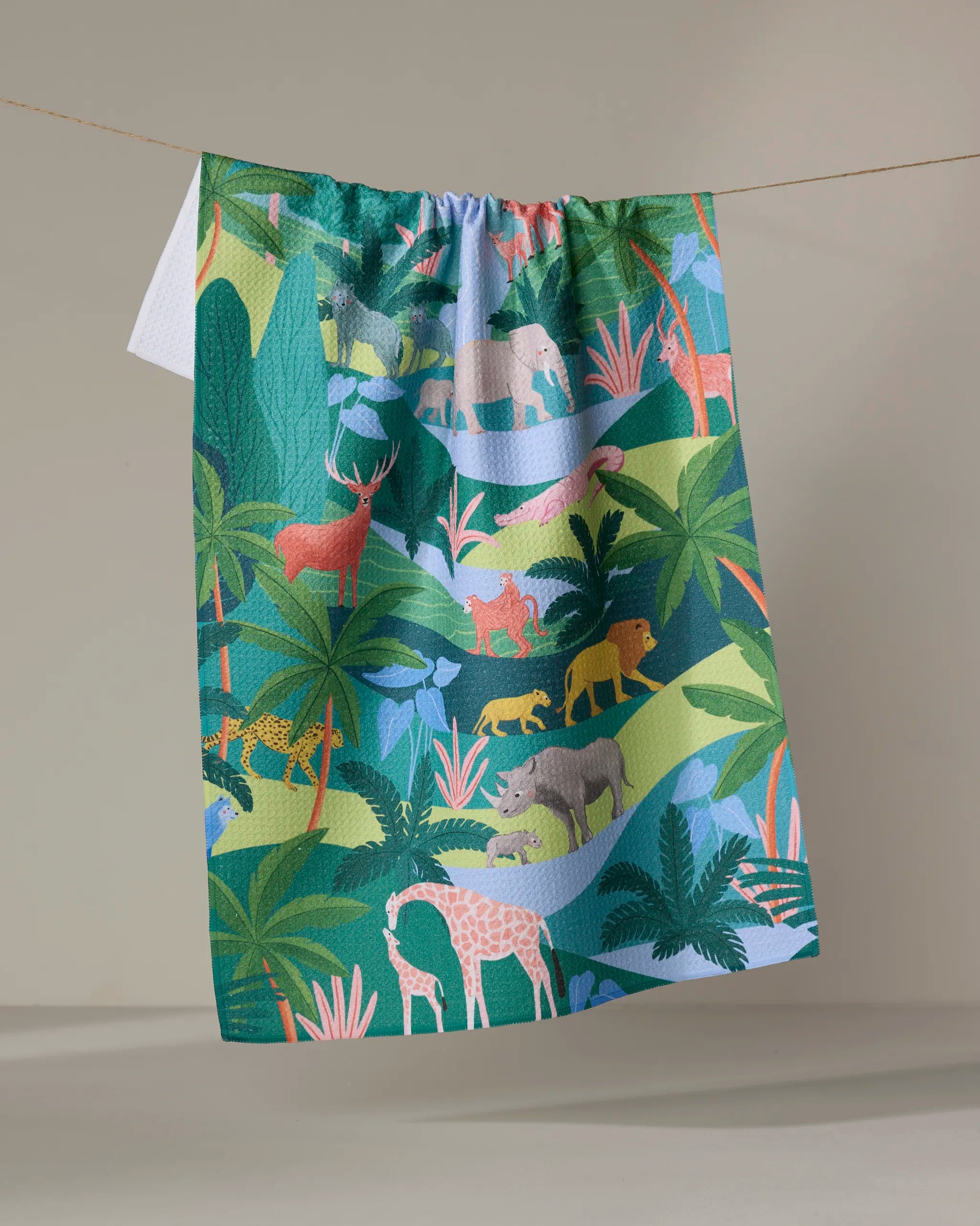 The "Rain Forest" tea towel by Ceyda Alasar, made from recycled materials and decorated with wild animal and tropical plant illustrations, hangs on a clothesline against a neutral background.