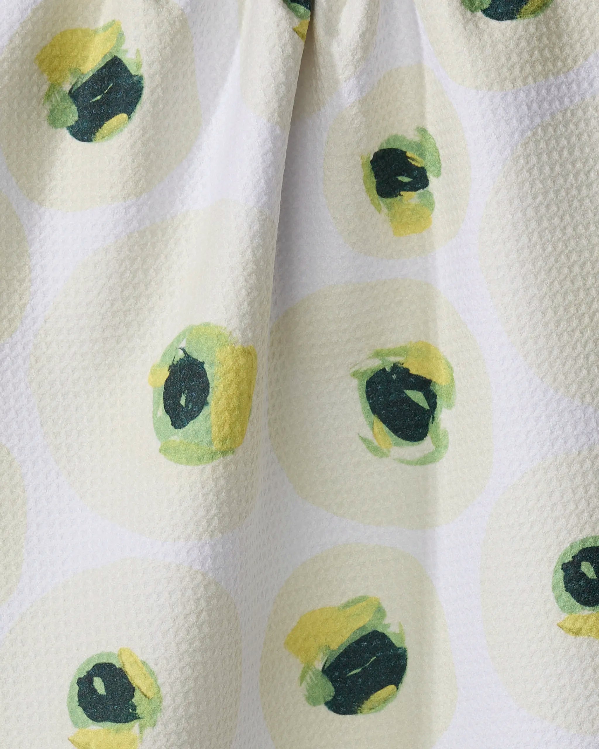 Close-up of the Ranunculus Eyes tea towel by Ania Zwara Art, made from recycled materials. Features textured white fabric with abstract green and yellow circles on a light yellow background. The ultra-absorbent towel is gently draped.