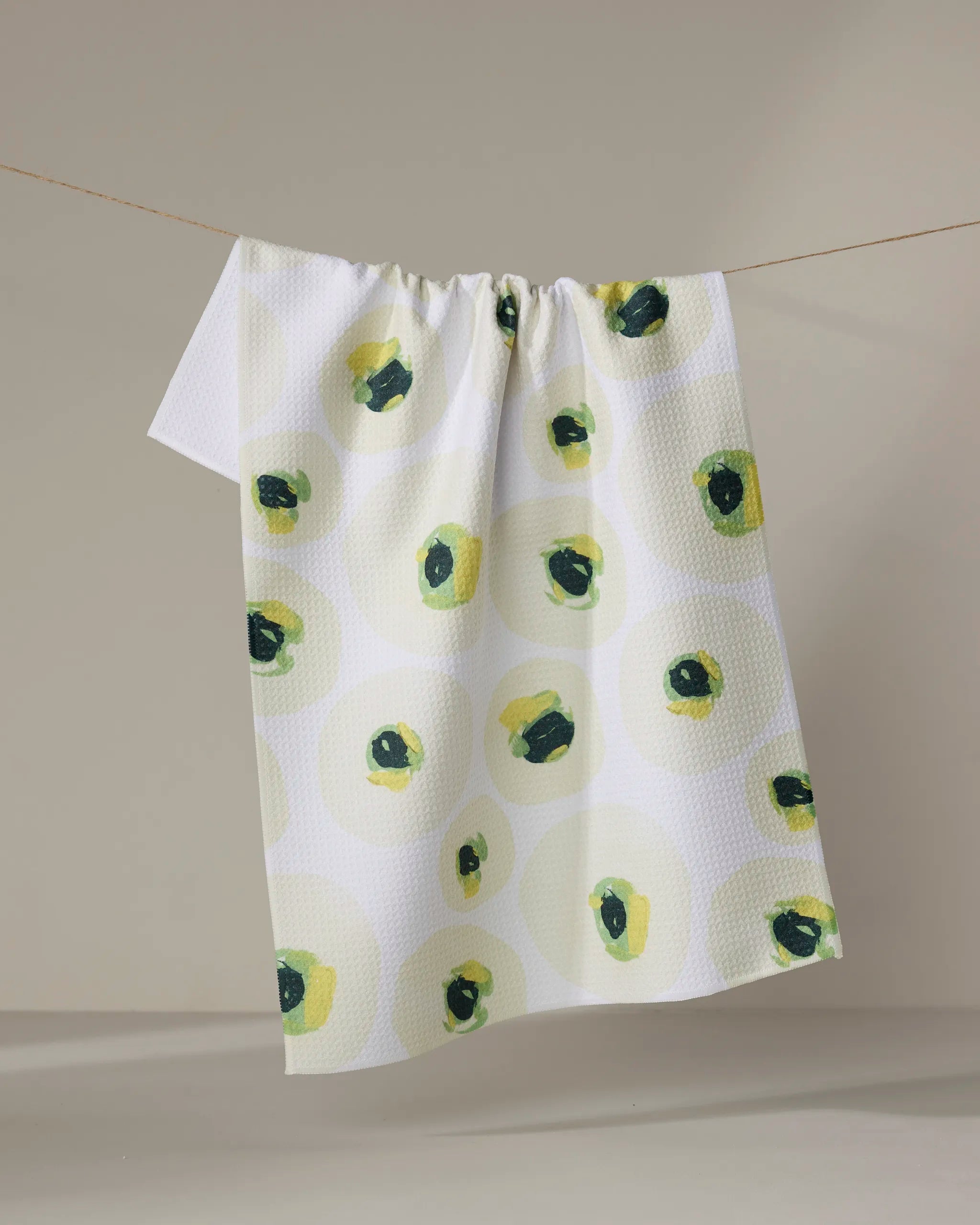 The "Ranunculus Eyes" tea towel by Ania Zwara Art, made from recycled materials, hangs on a line showcasing a textured surface and an abstract pattern of green and yellow circles.