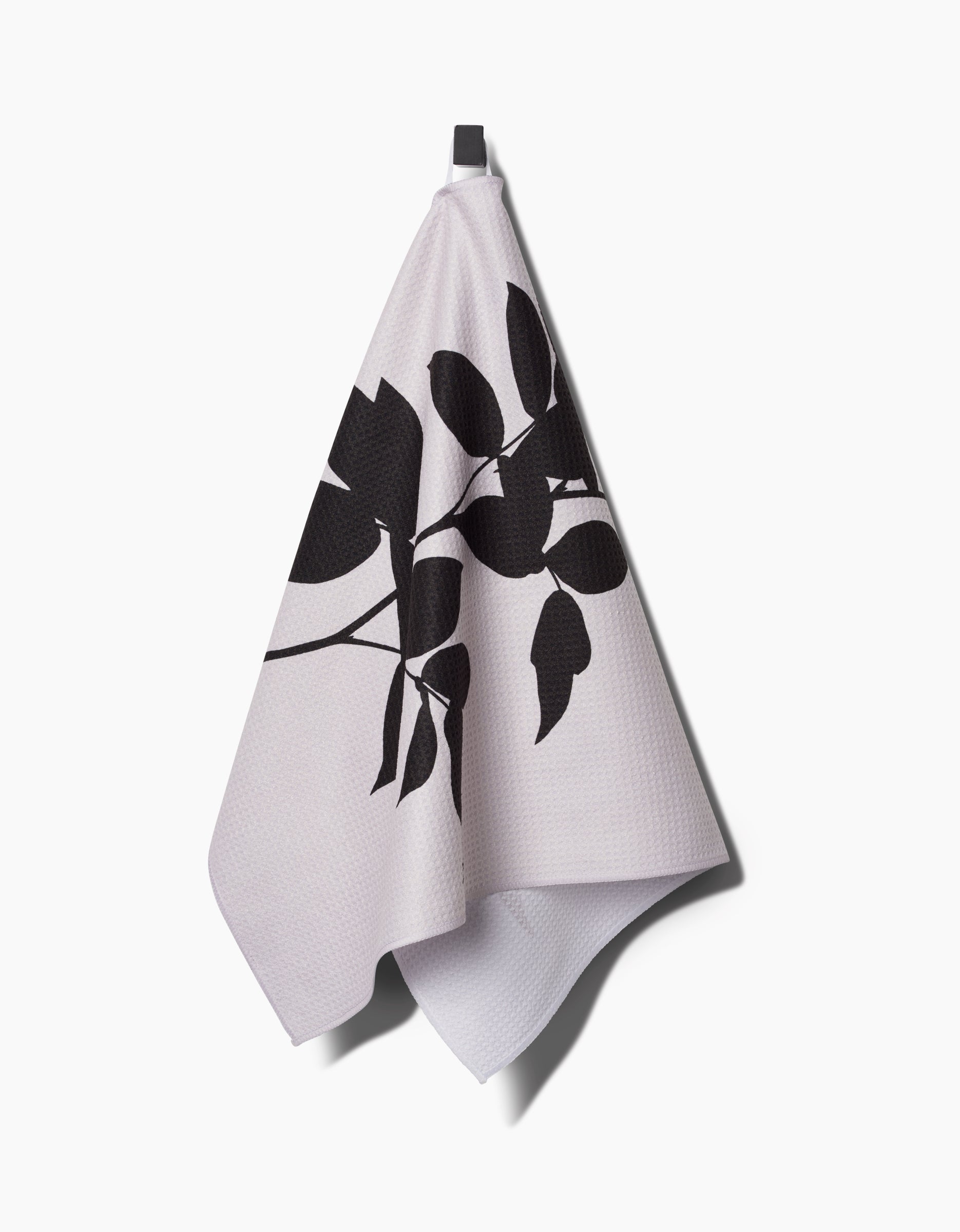 A light-colored PrintedPlant Raspberry Tea Towel made from Geoweave™ fabric with a black botanical leaf pattern hangs from a hook against a white background.