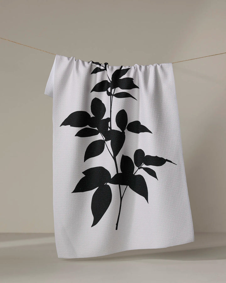 A PrintedPlant "Raspberry" white tea towel featuring a bold black botanical silhouette hangs on a thin clothesline against a neutral background.