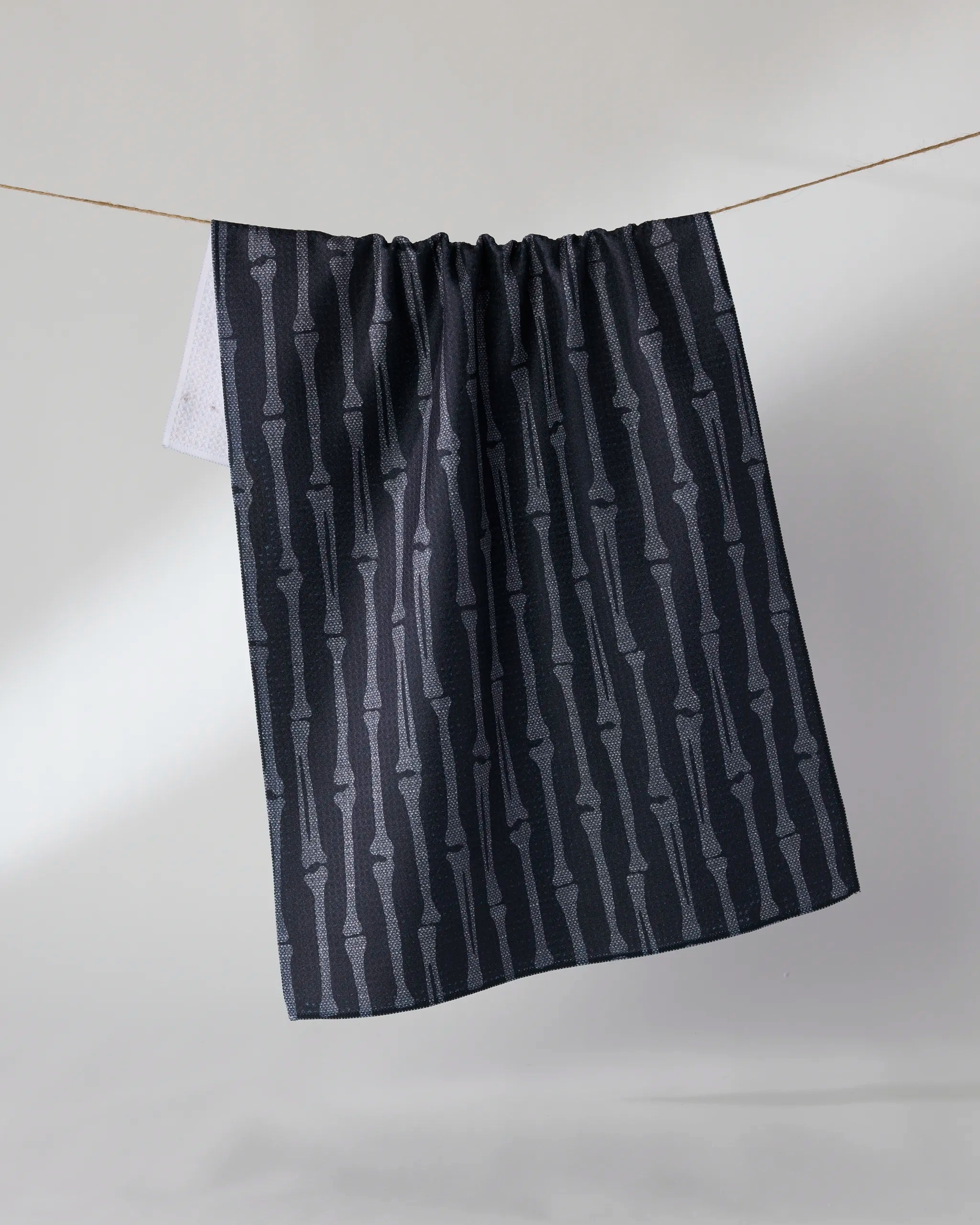 Geometry’s Rattler tea towel, featuring a subtle vertical bone pattern in dark hues, hangs on a clothesline against a light background.