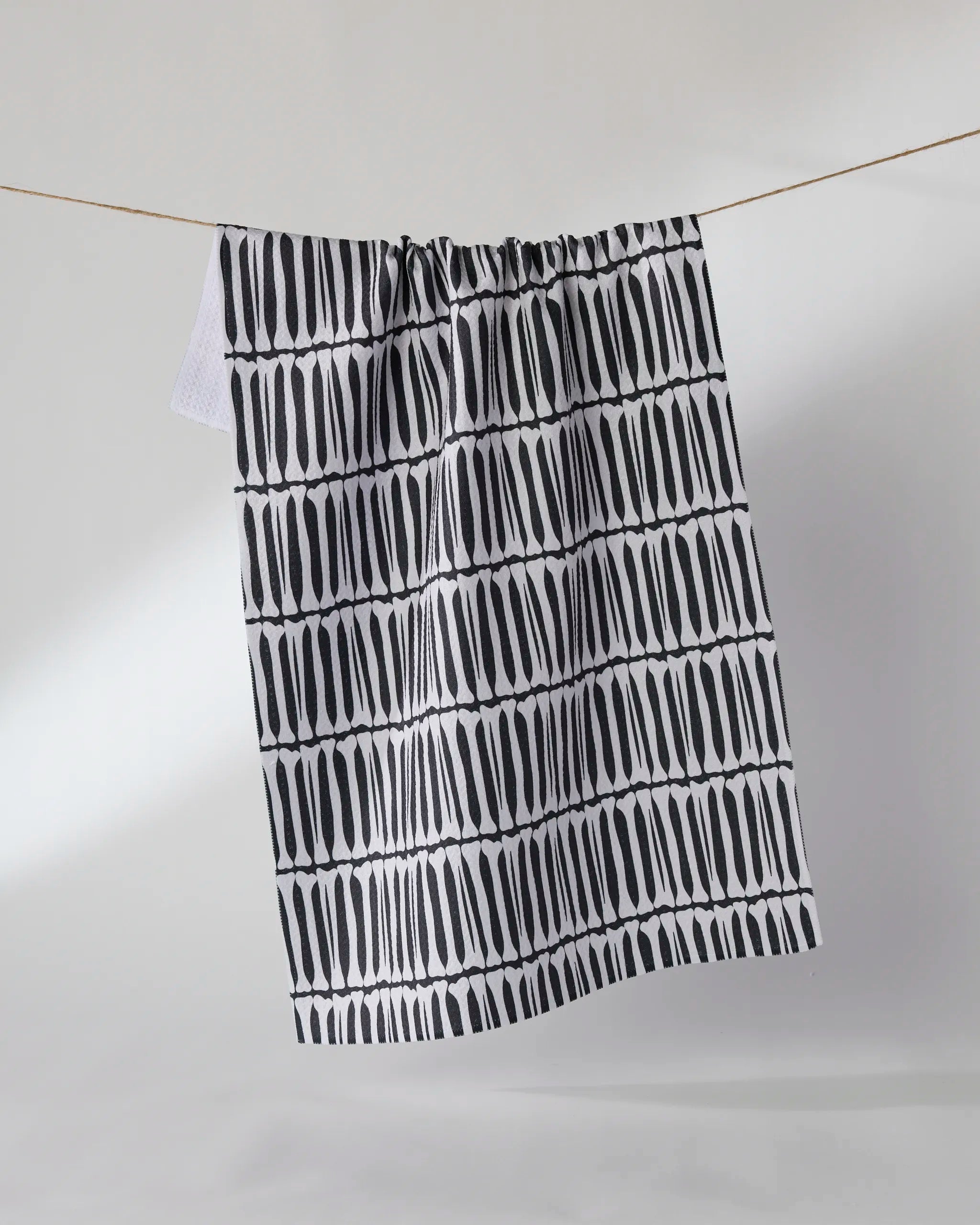 The Geometry Rattler tea towel, featuring hand-drawn black vertical stripes on white, hangs against a light background. Crafted from recycled materials, this ultra-absorbent towel blends stylish design with sustainability.