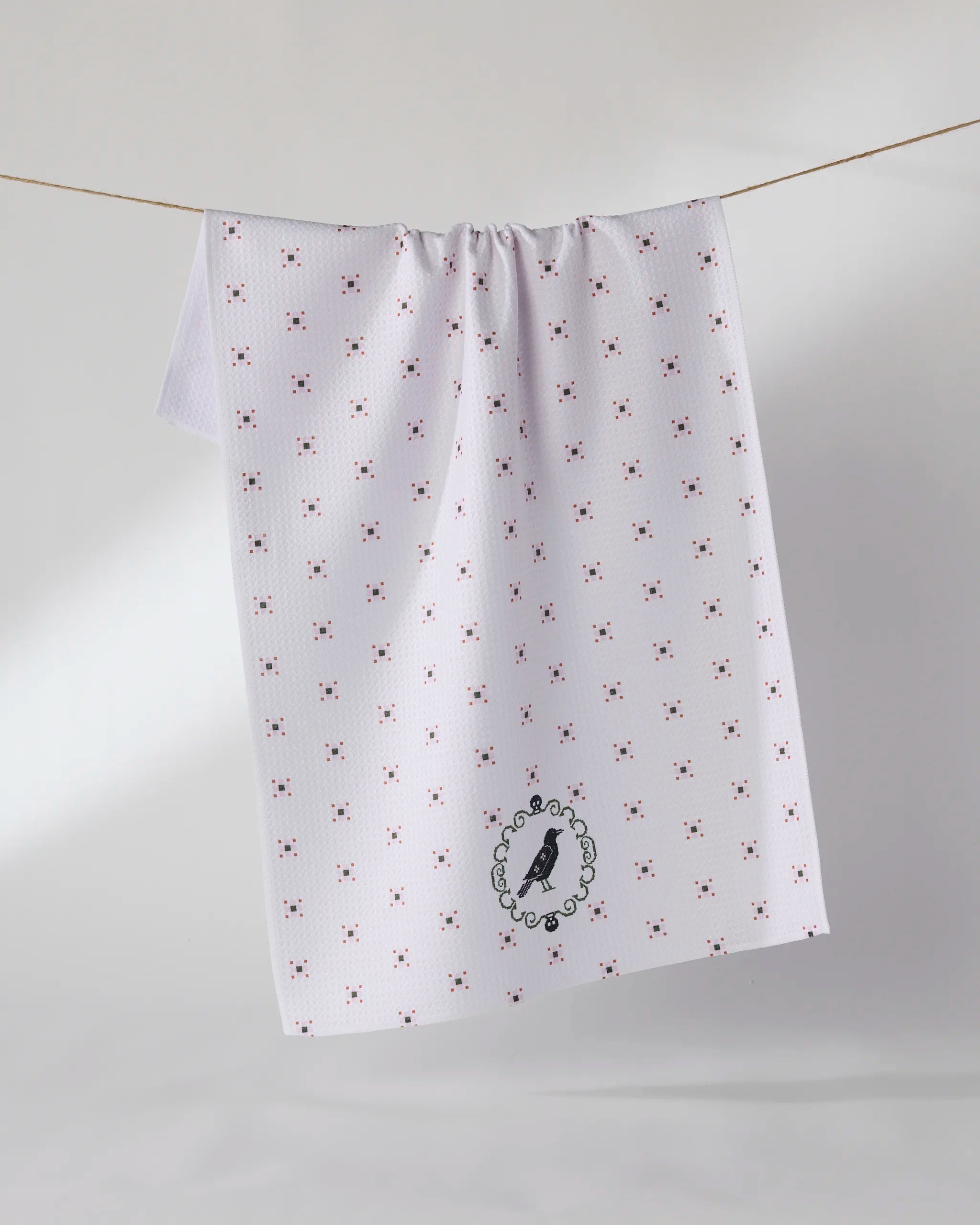 The Raven Bloom tea towel by Geometry features a white base with red and blue cross patterns, a black bird in a decorative oval near the bottom, and is crafted from recycled materials. A minimal background highlights its striking design.