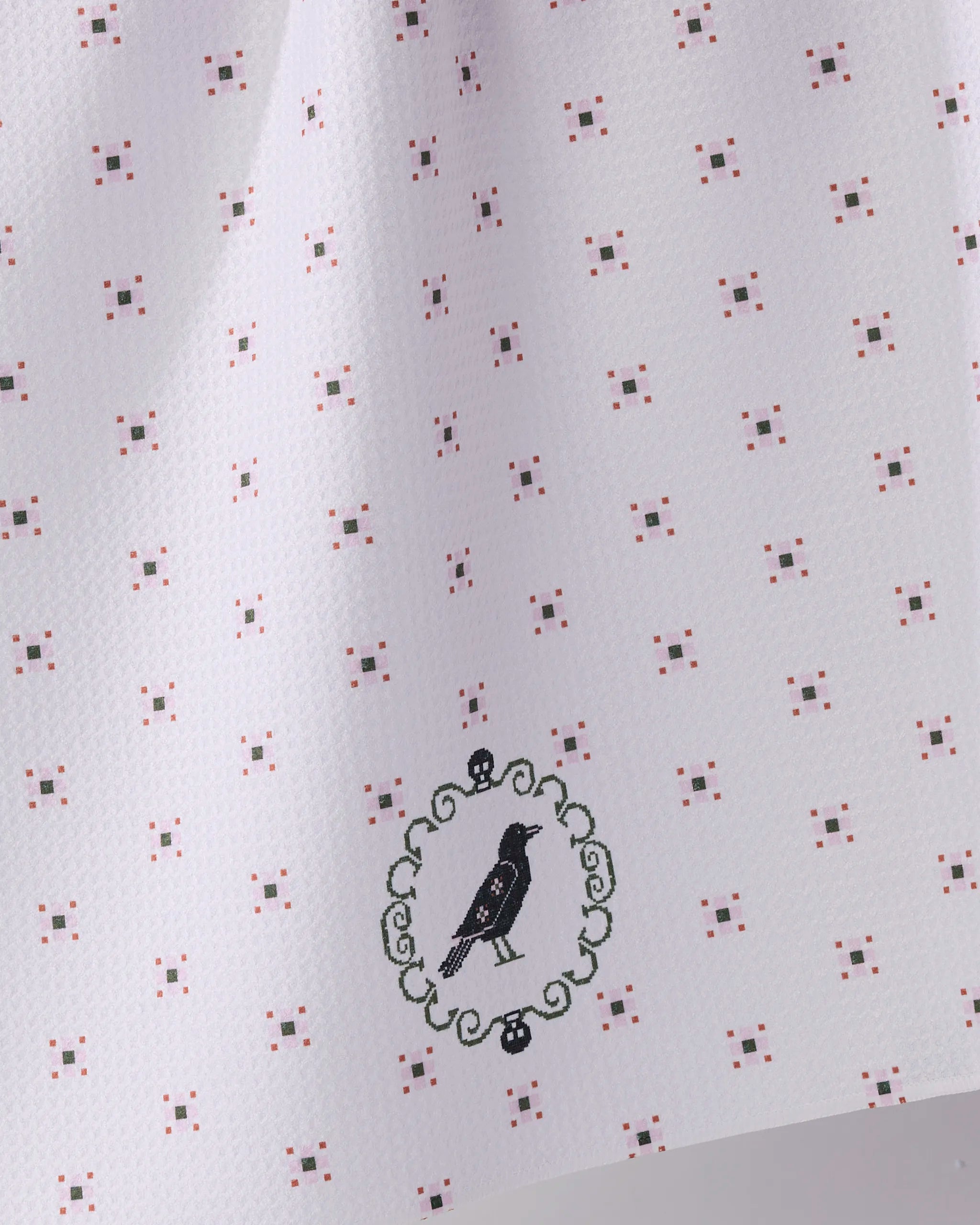 The Geometry Raven Bloom tea towel features a white textured design with a subtle grid of pink and brown dots, plus an embroidered black bird in a green oval frame. Made from quick-drying recycled materials.