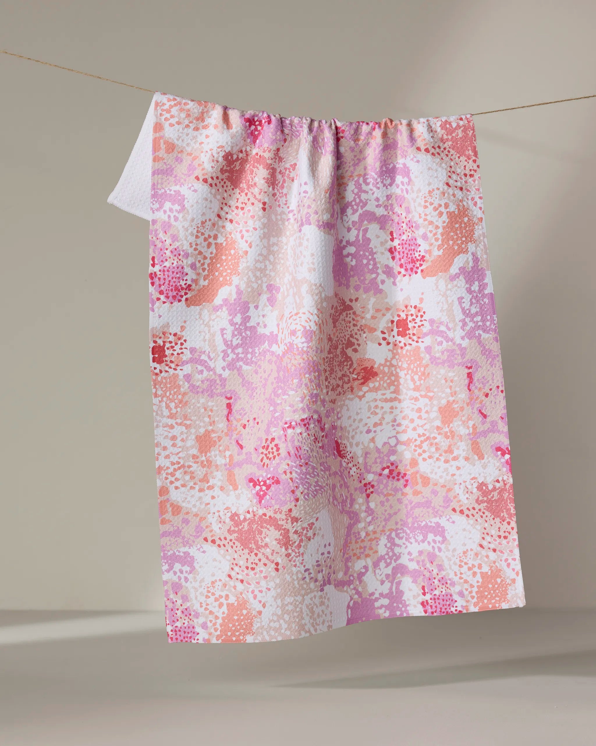 A rectangular recycled kitchen towel, Rawhide by Elisabeth Wing, featuring an abstract pink, peach, and white pattern hangs over a clothesline against a neutral background.