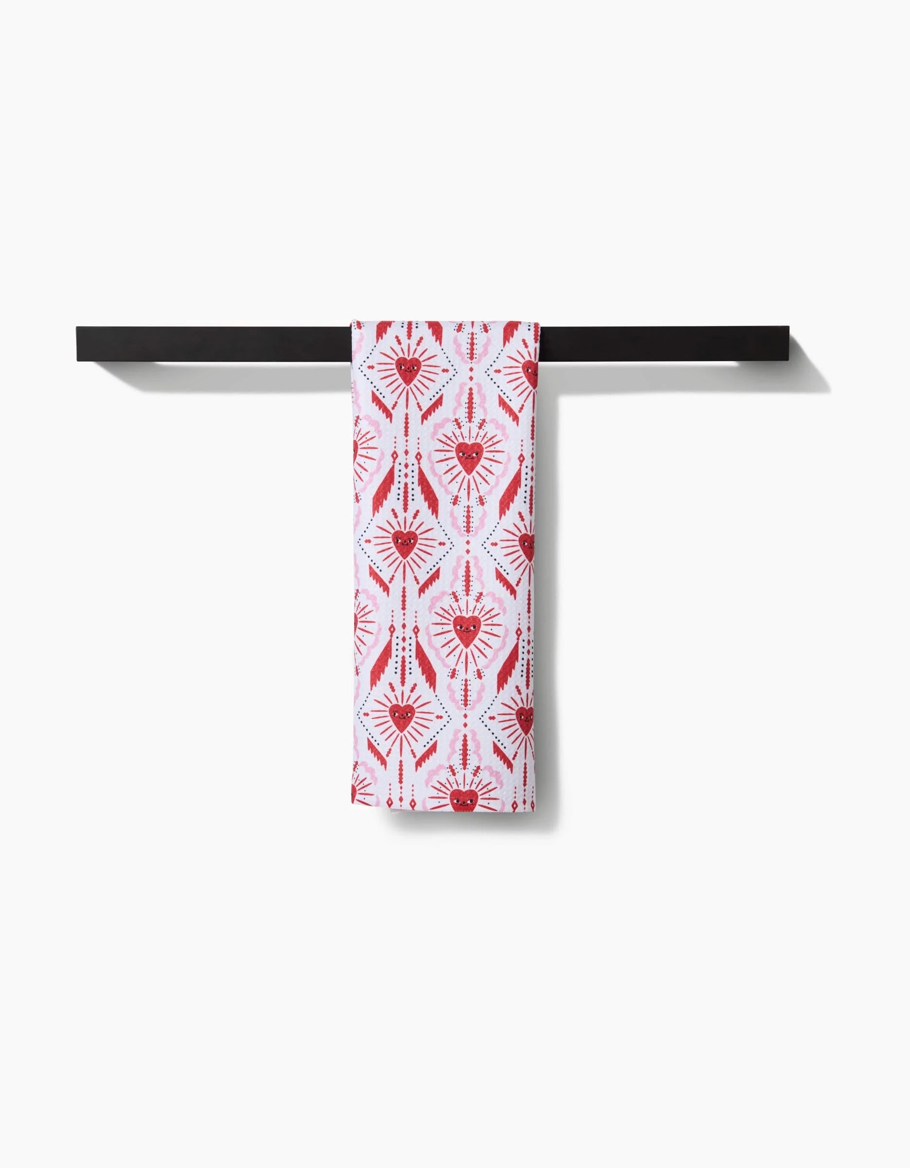 The Ray of Love recycled kitchen towel by Roxy Marj, featuring red geometric and heart patterns, is draped over a simple black horizontal towel bar against a white background.
