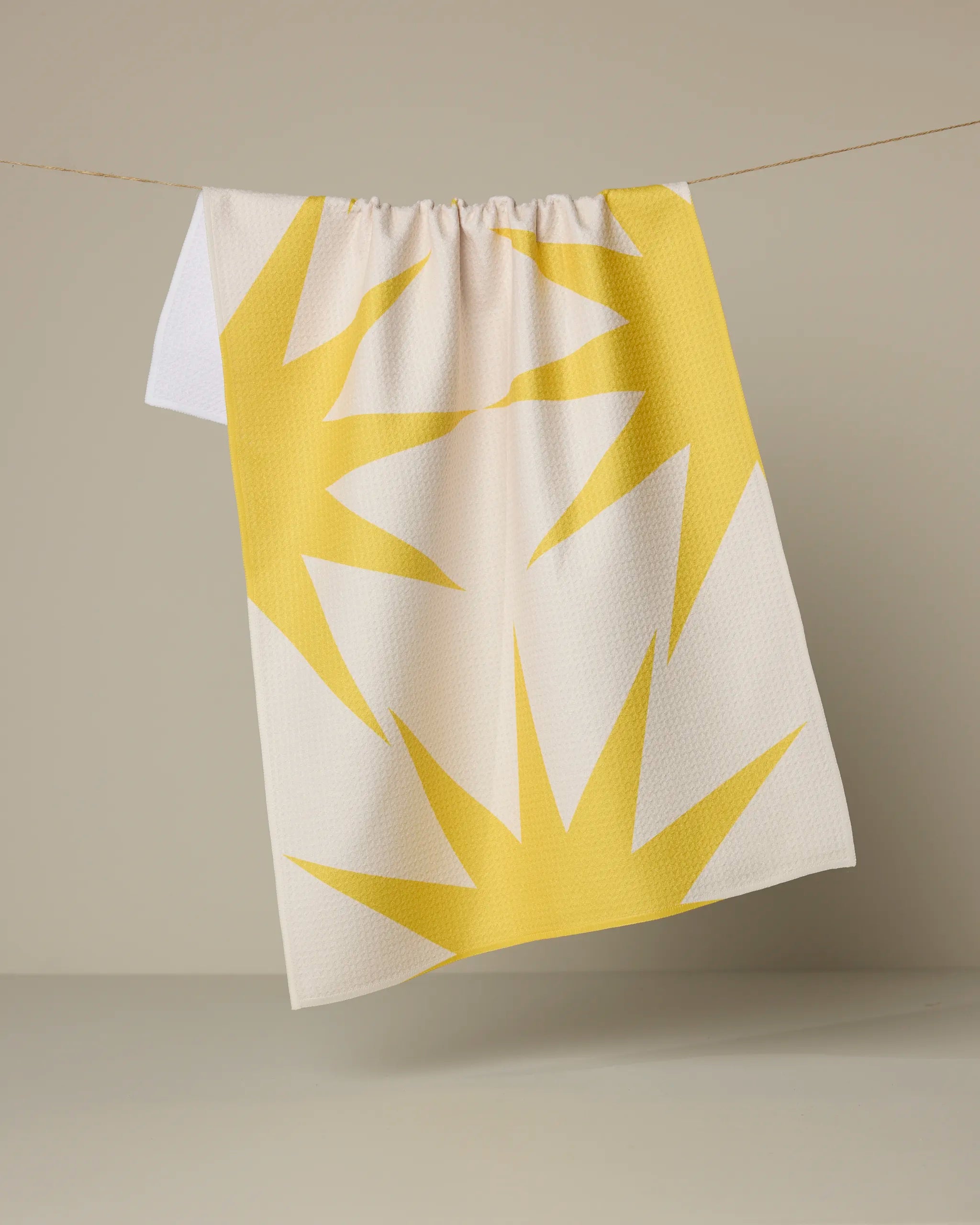 The Ray tea towel by Geometry features a bold yellow sunburst pattern on white, made from recycled materials, and hangs on a thin string against a neutral background.