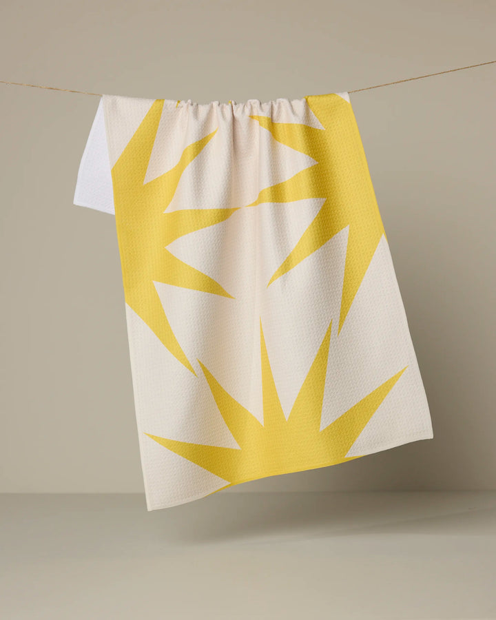 The Ray tea towel by Geometry features a bold yellow sunburst pattern on white, made from recycled materials, and hangs on a thin string against a neutral background.