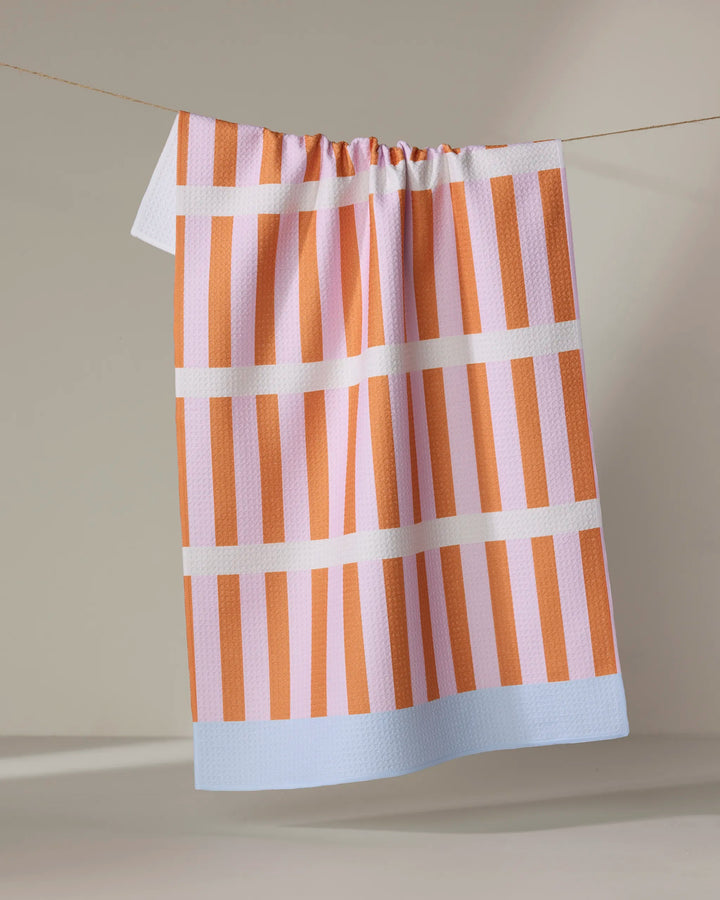 The "Red Glow" tea towel by Happy Hemera, featuring pink and orange stripes and a light blue border, is made from recycled materials and hangs on a clothesline against a neutral backdrop.