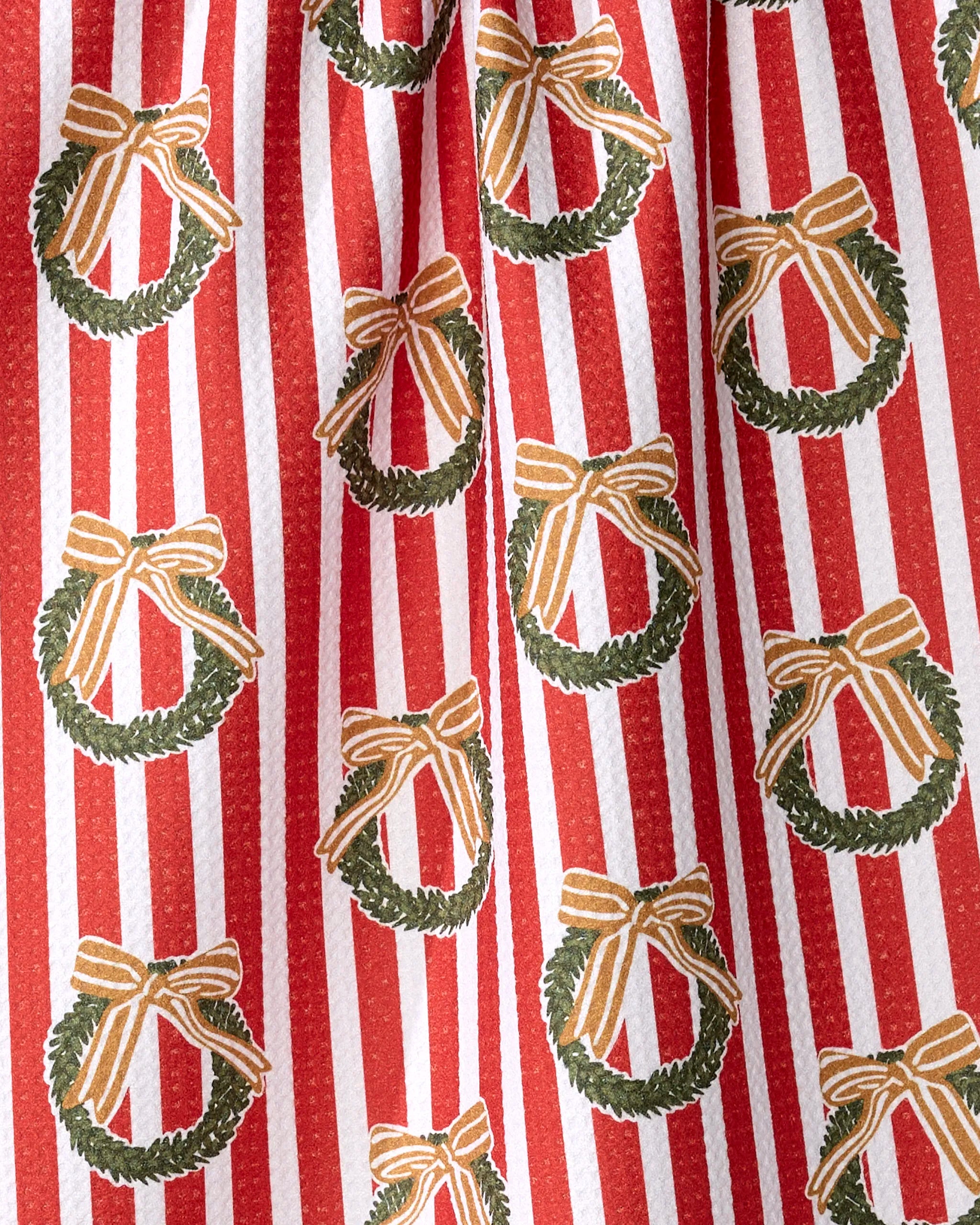 The Red Wreaths tea towel by Charlie Rowan Designs features red and white stripes with green wreaths and gold bows, perfect for holiday decor. Made from recycled materials.