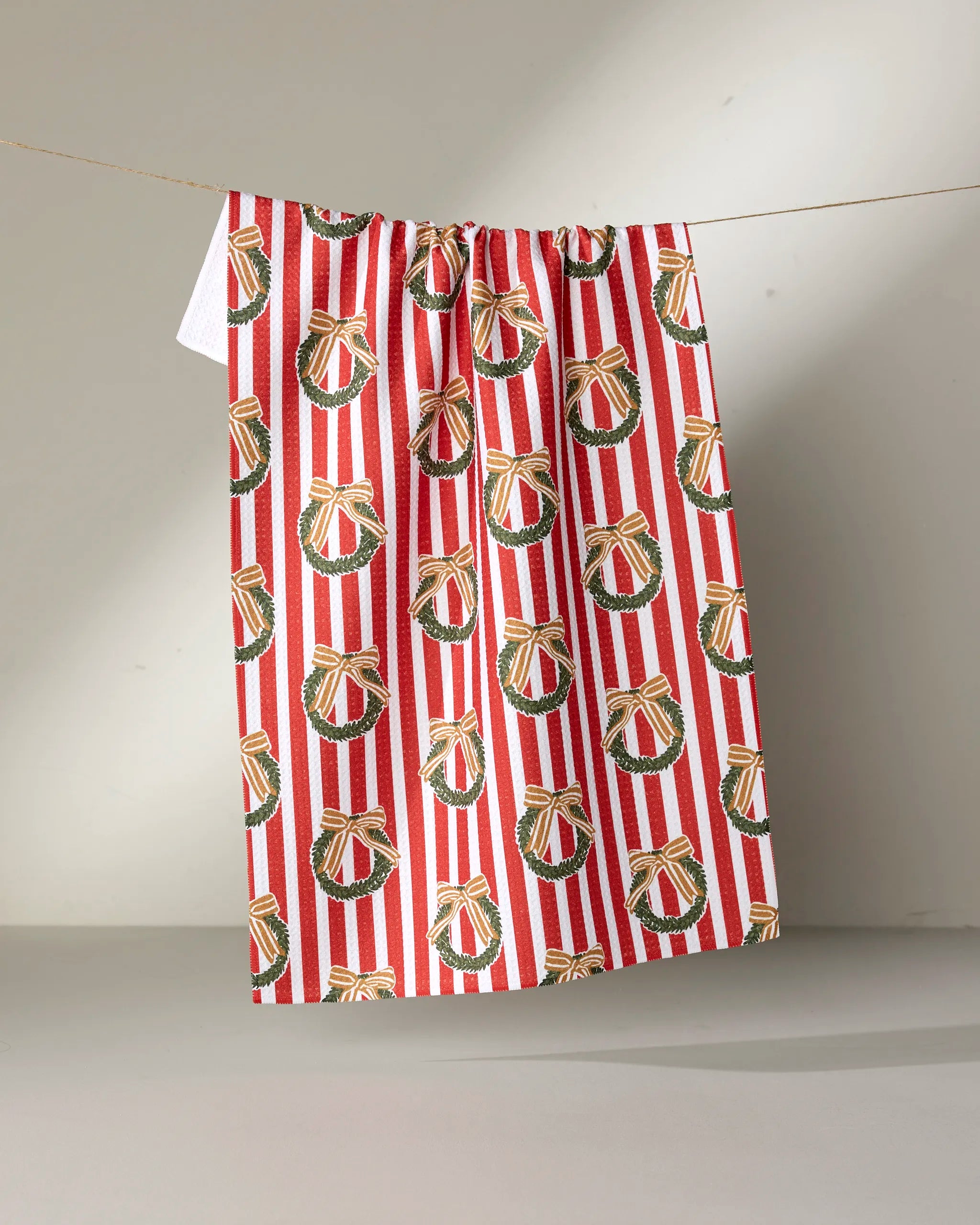 The "Red Wreaths" kitchen tea towel by Charlie Rowan Designs features green wreaths with gold bows and red ornaments on red and white stripes, and is crafted from recycled materials.