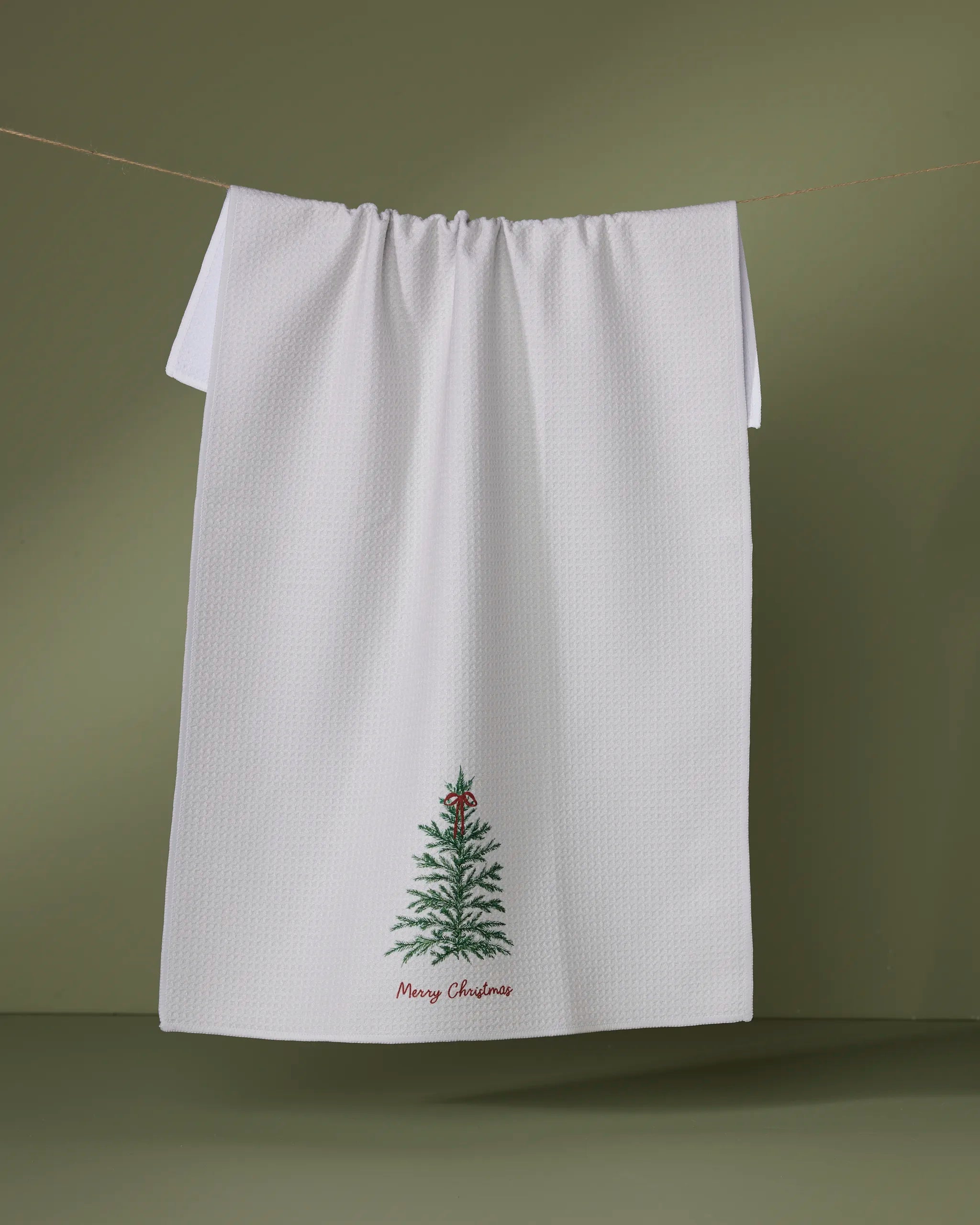 A Geometry Redford white recycled kitchen towel featuring a small embroidered Christmas tree and "Merry Christmas" hangs on a clothesline against a green background.