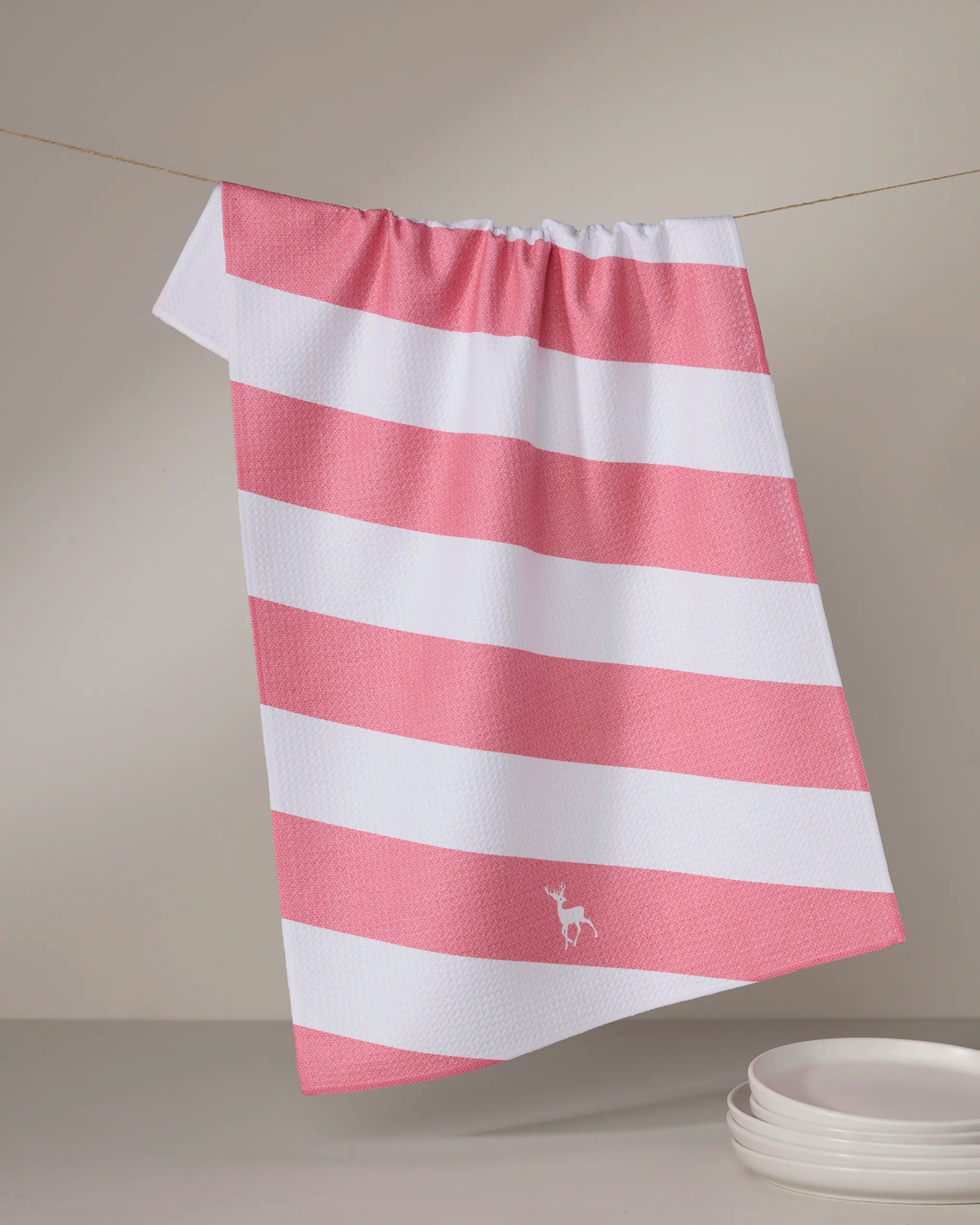 The Geometry "Reindeer Run" tea towel, made from recycled materials, displays pink and white stripes with a small deer logo in the corner. It hangs against a neutral background, next to a stack of white plates.