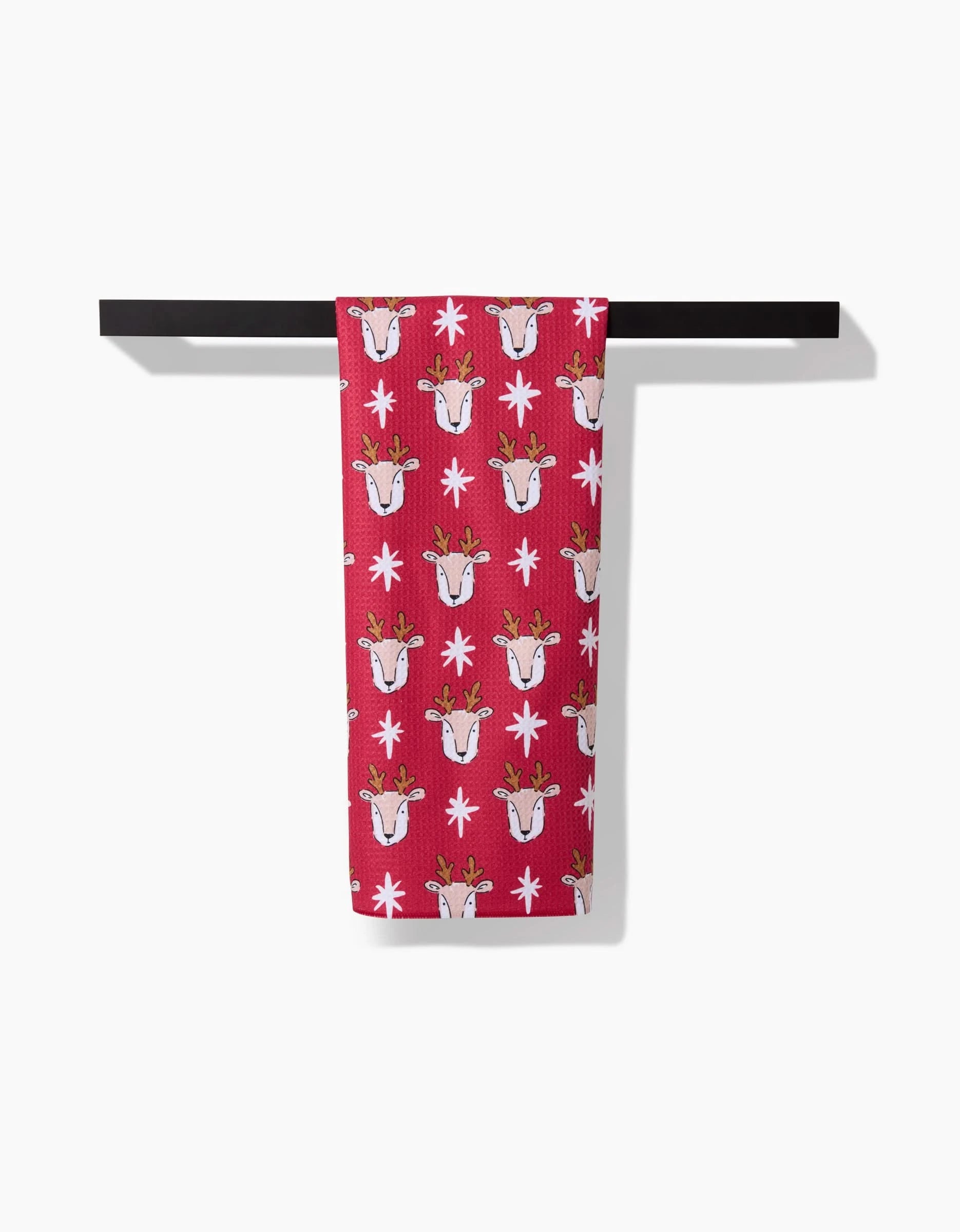 The Reindeer Star kitchen towel by Hufton Studio features a repeating pattern of cartoon reindeer faces and white snowflakes in red, shown hanging on a horizontal black bar against a white background.