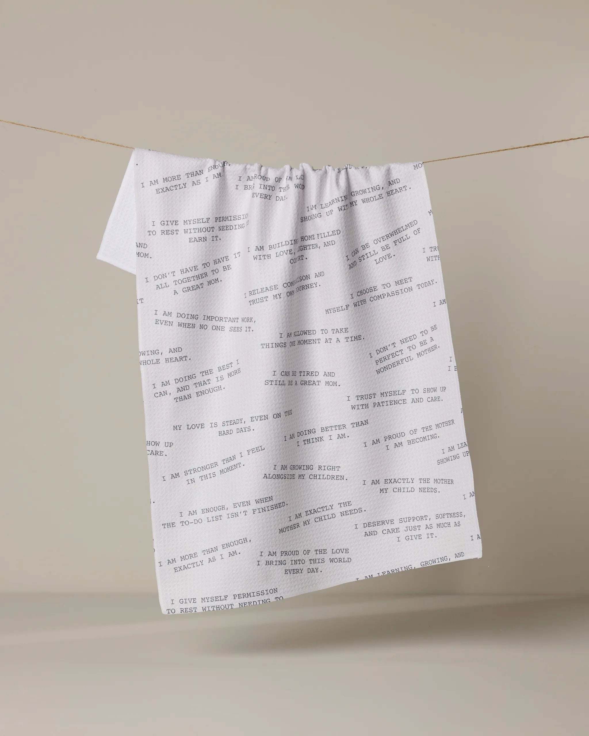 The Geometry "Reminders for Mom" kitchen towel, white with black affirmations and positive messages, hangs on a clothesline against a neutral beige background.
