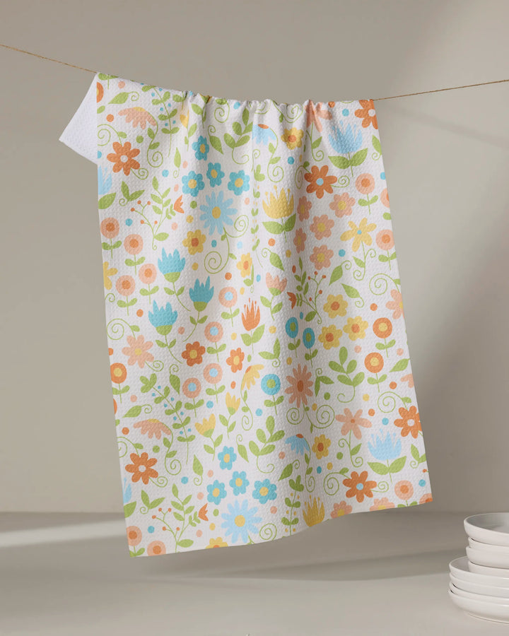 The Mindfulnice Remindful Rememberings tea towel, featuring a vibrant floral pattern in orange, blue, pink, and green on white, hangs on a clothesline with stacked white plates nearby.