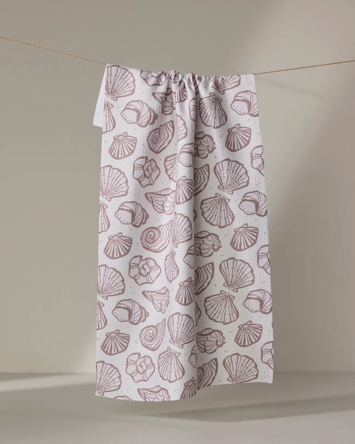The Retro Coastal Seashells in Neutral tea towel by Teri the Butter, made from recycled materials, features an all-over light brown seashell outline print on white and hangs against a plain background.