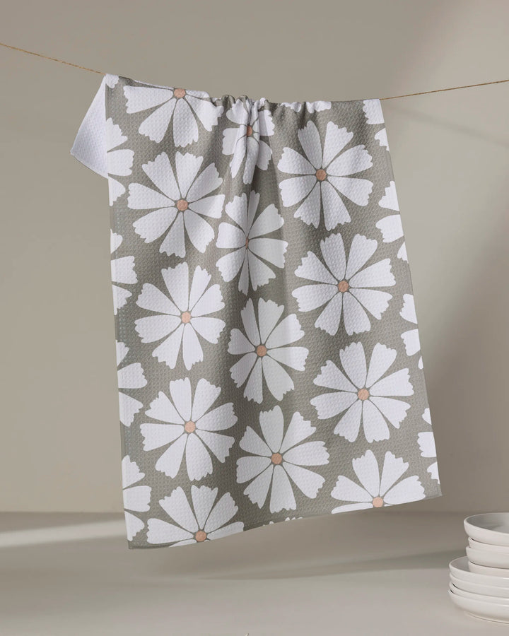 The Hufton Studio Retro Daisy tea towel, made from recycled materials and featuring large white flowers with brown centers, hangs on a line. A stack of white plates sits in the lower right corner against a neutral backdrop.