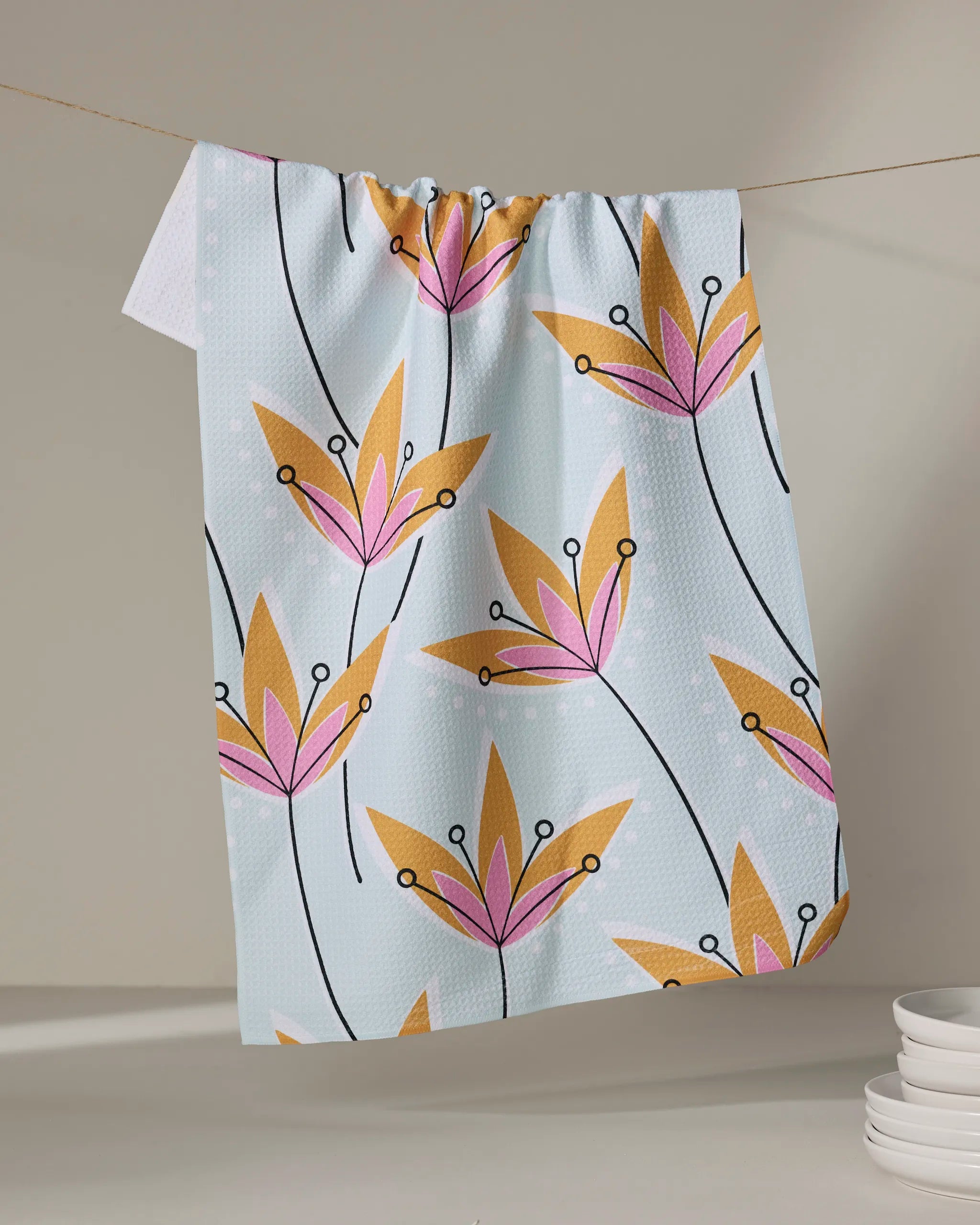 The Retro Floral tea towel by Julianne Haness, in light blue with pink and yellow flowers, dries quickly as it hangs indoors on a clothesline, while a stack of white plates sits in the lower right corner.