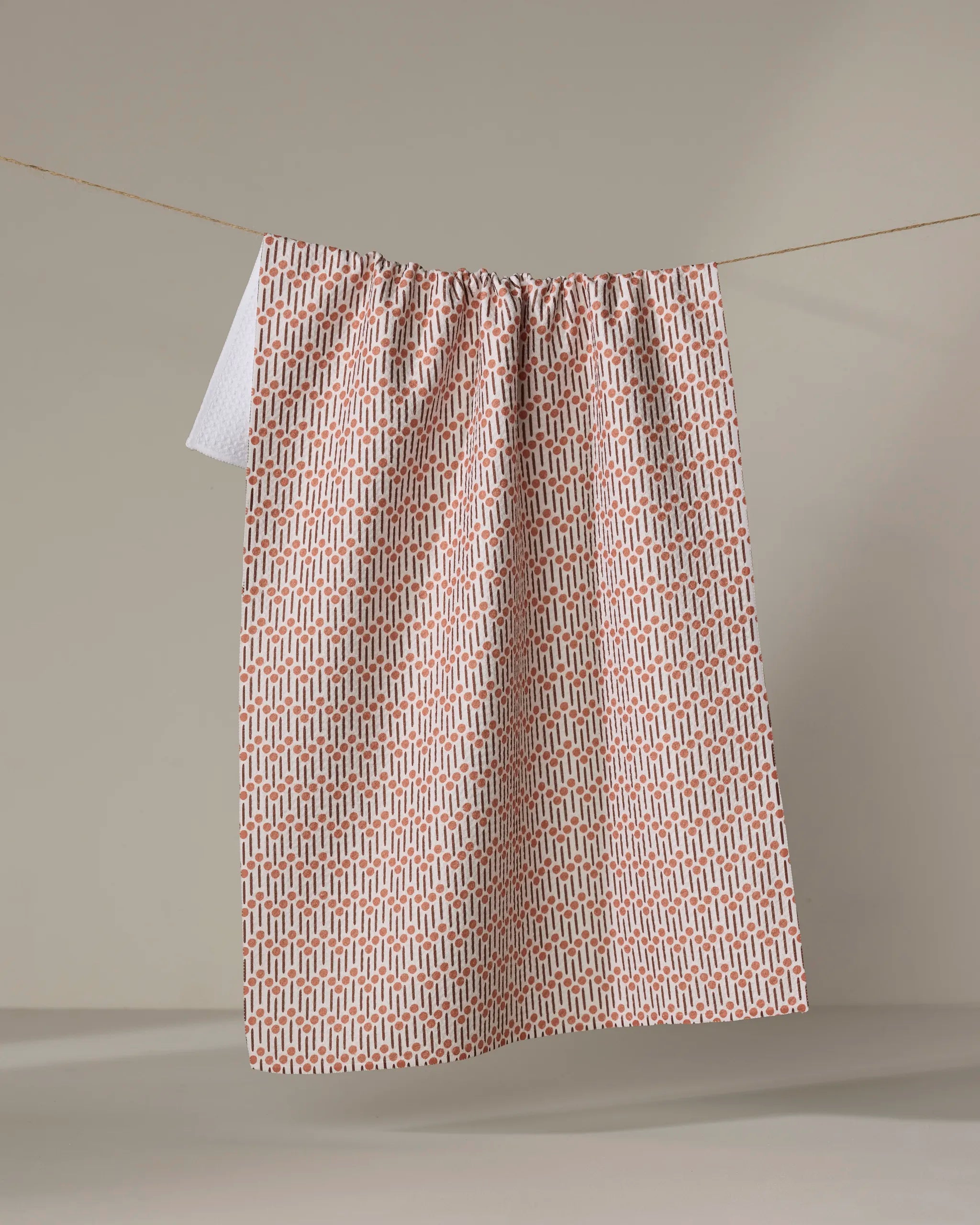 The Retro Geo tea towel by Teri the Butter, made from recycled materials and featuring a pink and white geometric pattern, hangs on a clothesline against a plain beige background.