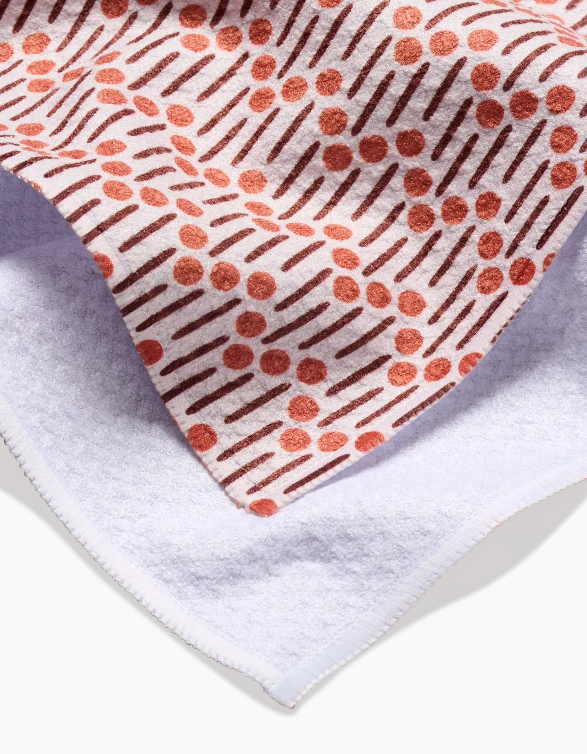 A close-up of a Teri the Butter Retro Geo tea towel shows its white terry cloth side and a patterned side with orange dots and lines on white, made from recycled materials. The patterned corner is folded over the plain side.
