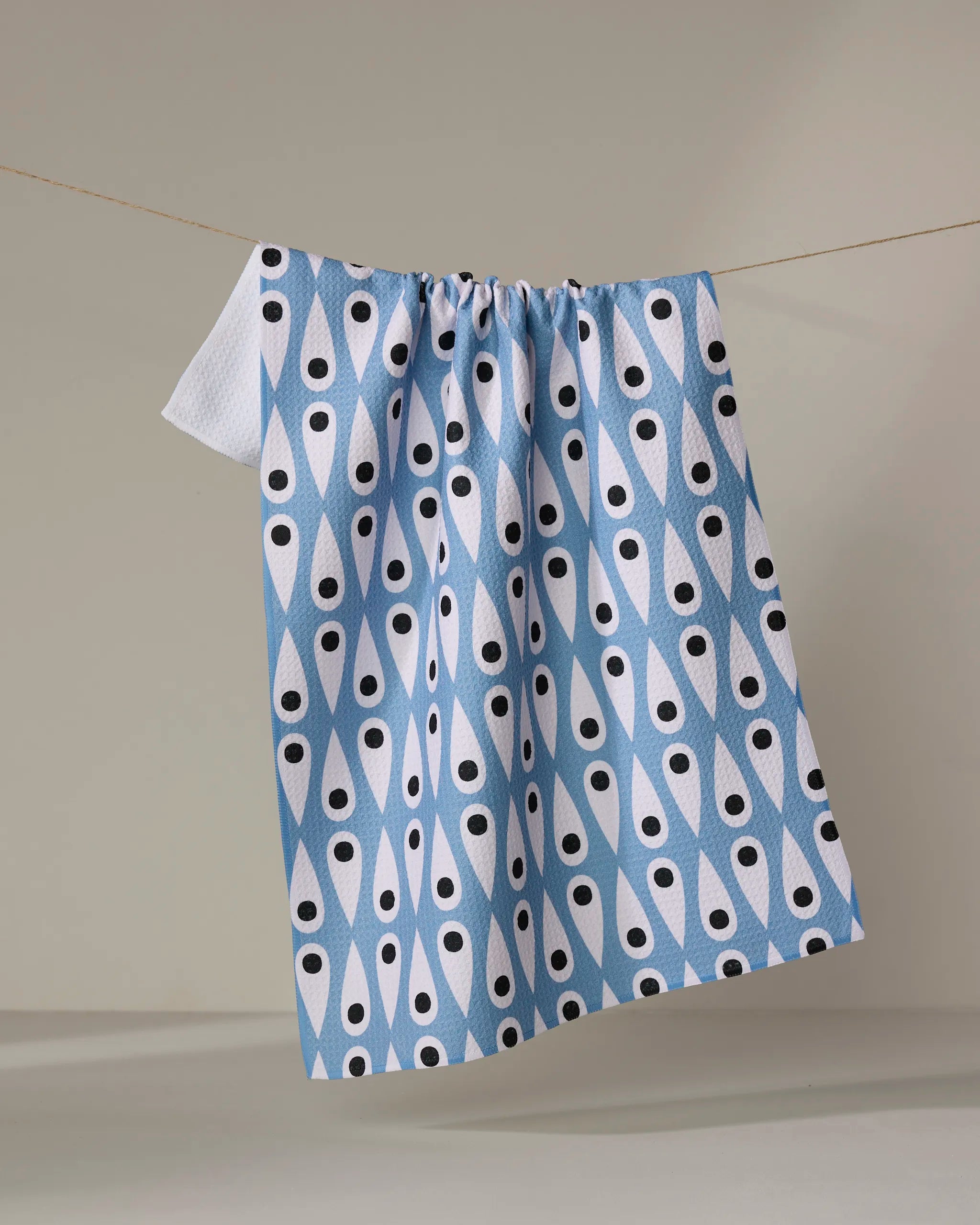 The Retro Rain tea towel by Anette Heiberg, featuring blue and white patterns with black teardrop shapes, hangs from a clothesline against a plain beige background.