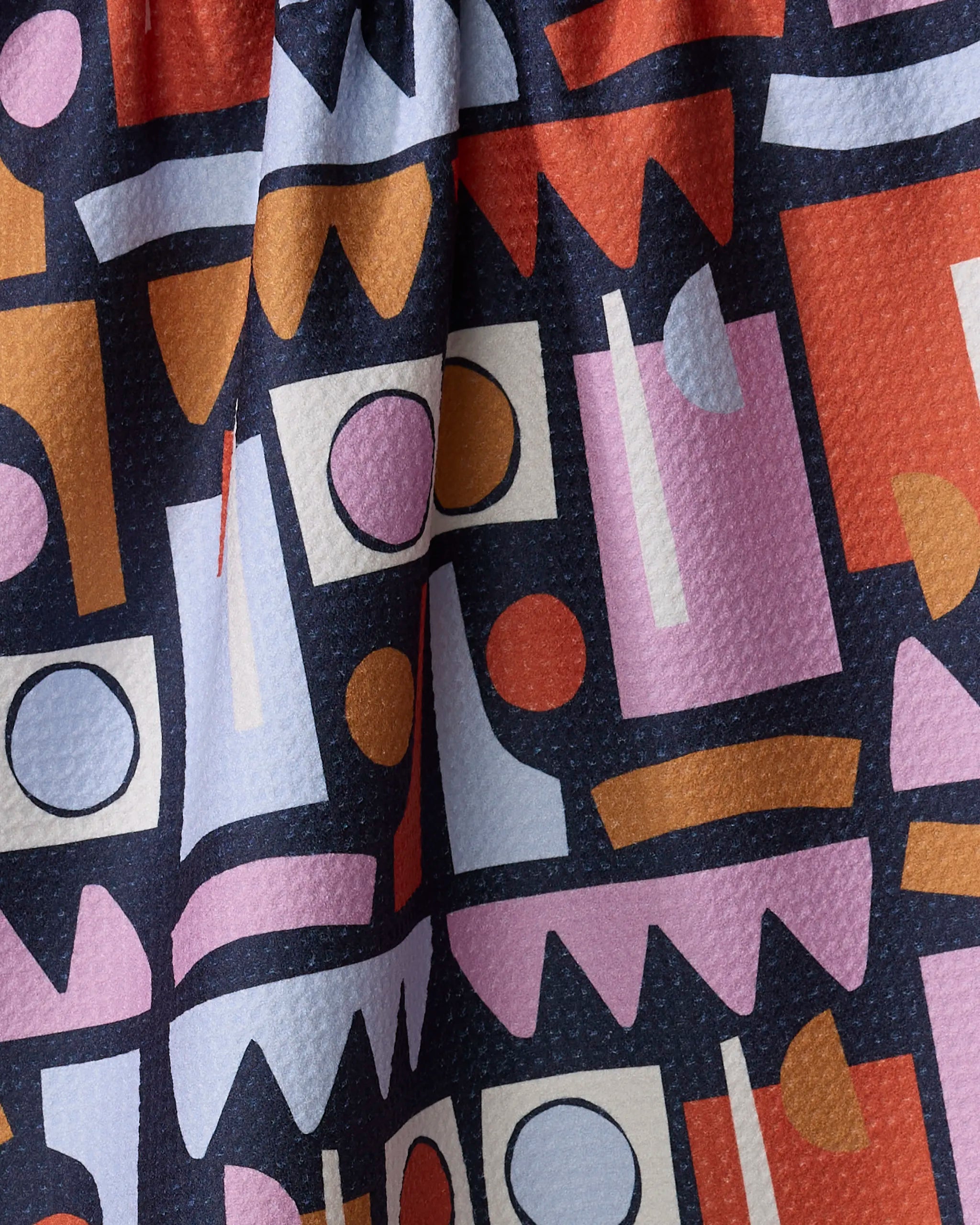 A close-up of the Retro Shaped Fiesta tea towel by Claire Iglesias, with a colorful geometric pattern—circles, rectangles, and abstract shapes in orange, red, pink, purple, and white on a dark background. The fabric appears slightly gathered.