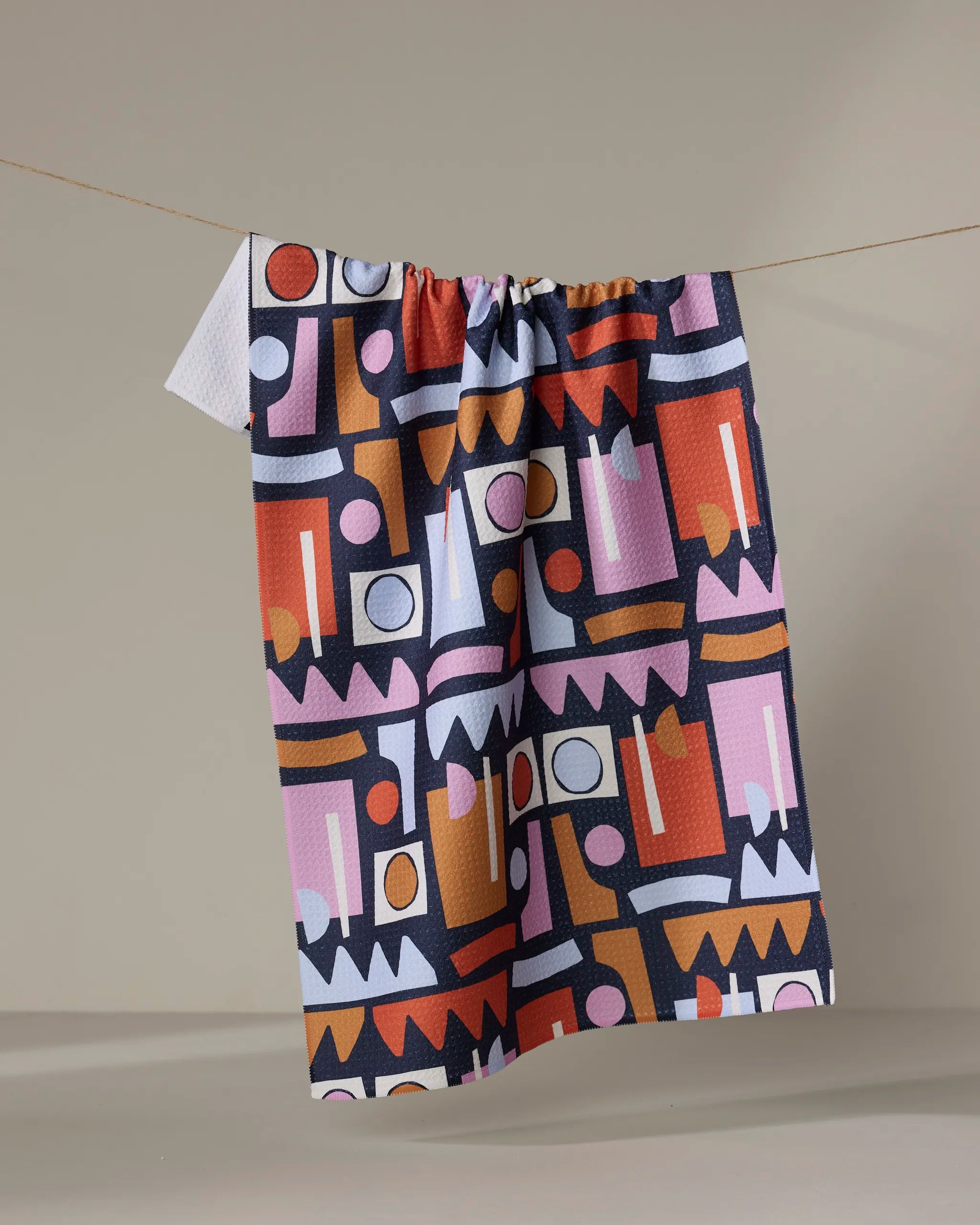 The Retro Shaped Fiesta tea towel by Claire Iglesias, featuring a bold geometric pattern in pink, orange, white, black, and red, hangs on a string against a plain beige background.