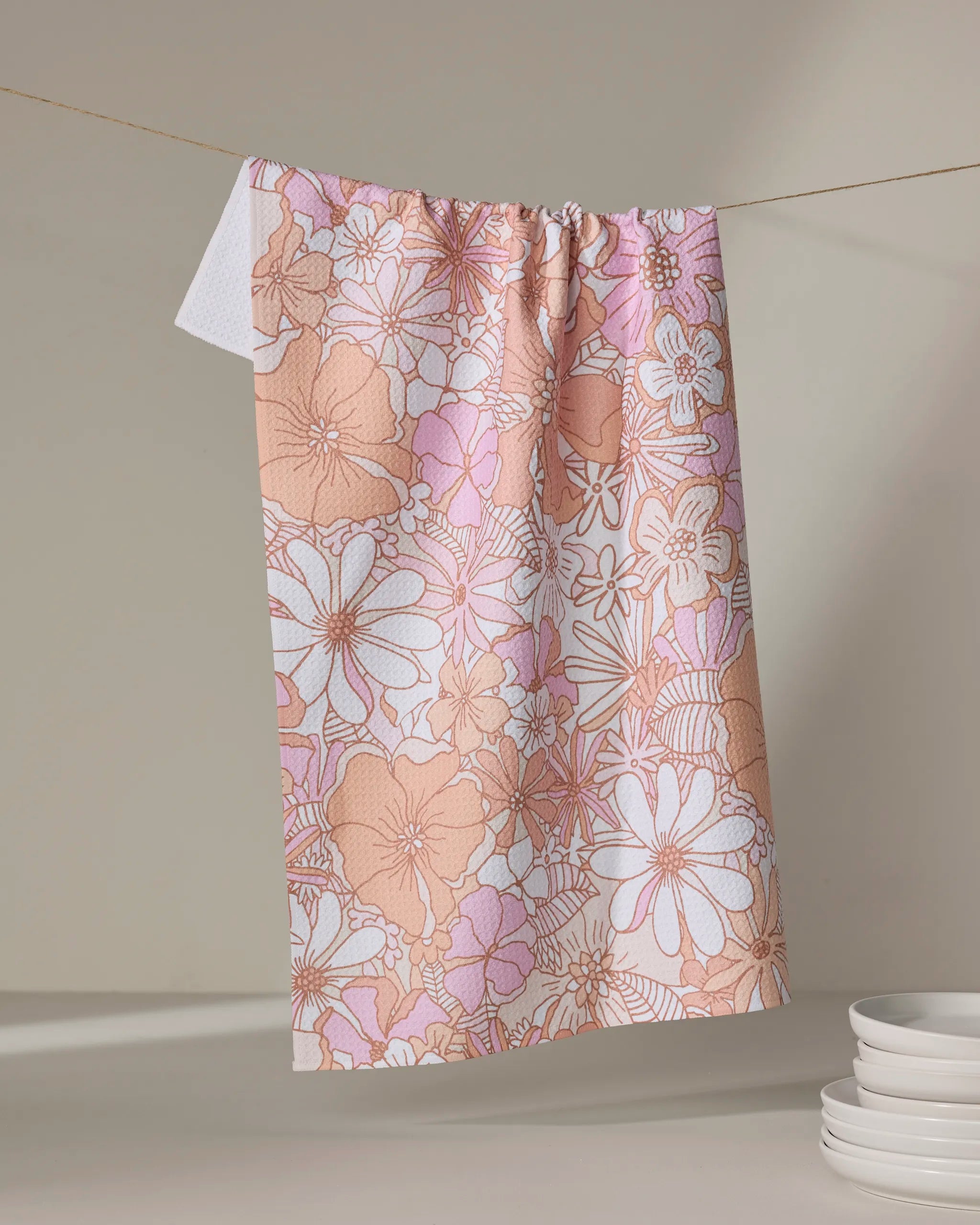 The Retro Vibes tea towel by Marina Solodka, featuring a pink and beige floral pattern and ultra-absorbent fabric, hangs on a clothesline against a neutral background beside a stack of white plates.