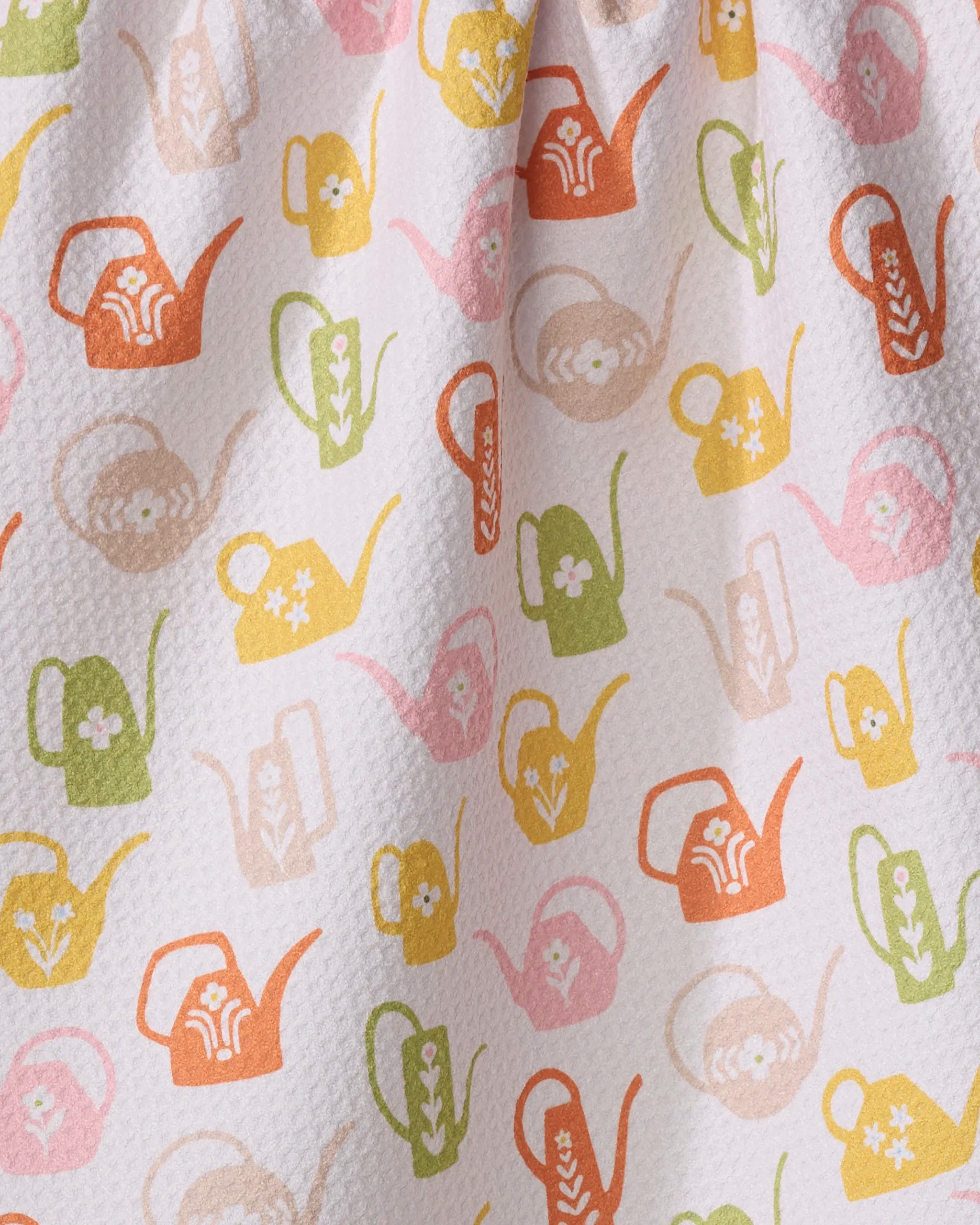 Kayla Ann’s Retro Watering Cans kitchen towel is textured, white, and made from recycled materials. It features a repeating pattern of watering cans in orange, green, yellow, and pink with unique floral designs.