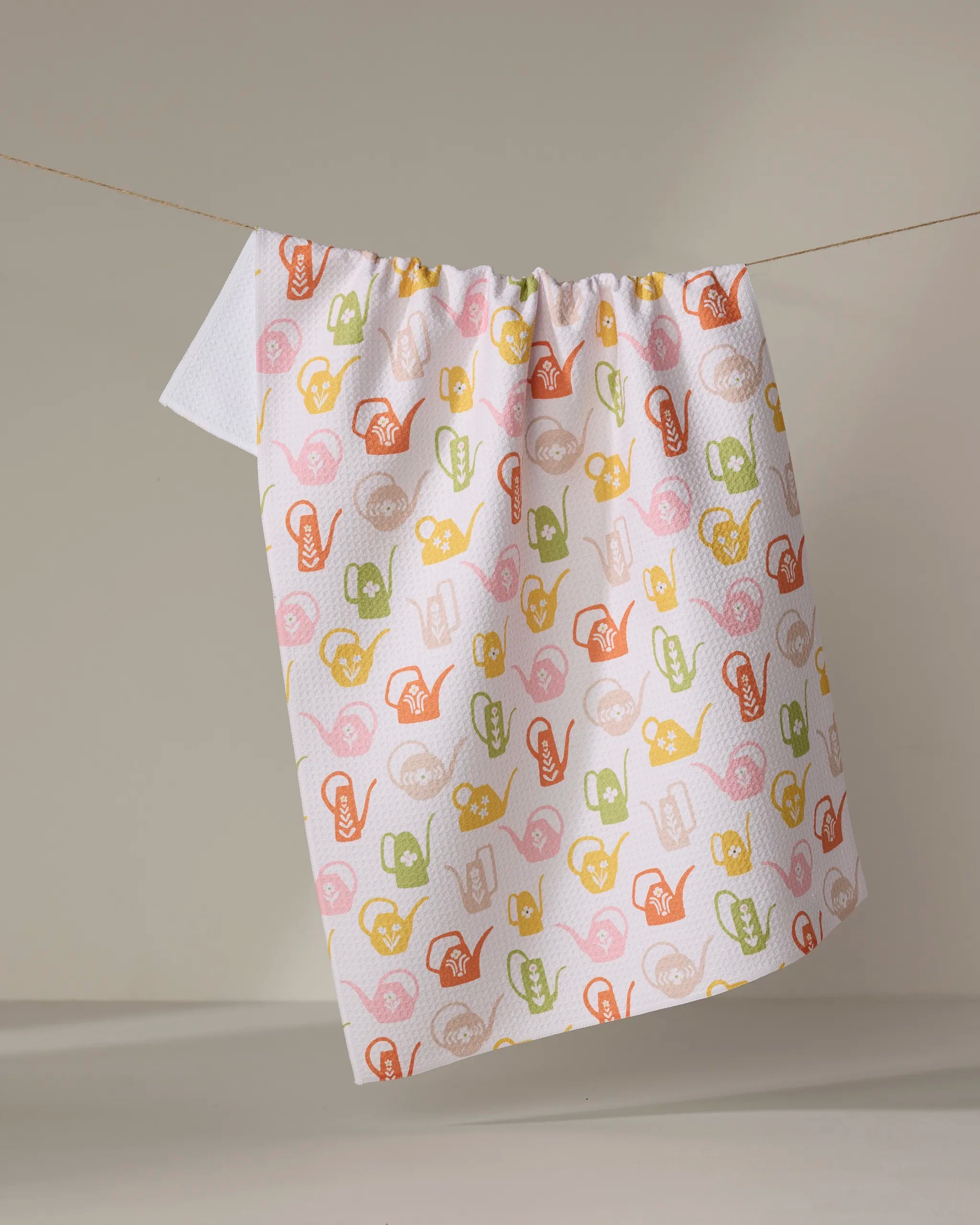 A white ultra-absorbent dish towel by Kayla Ann featuring a colorful retro watering cans pattern hangs on a clothesline against a plain, light background.