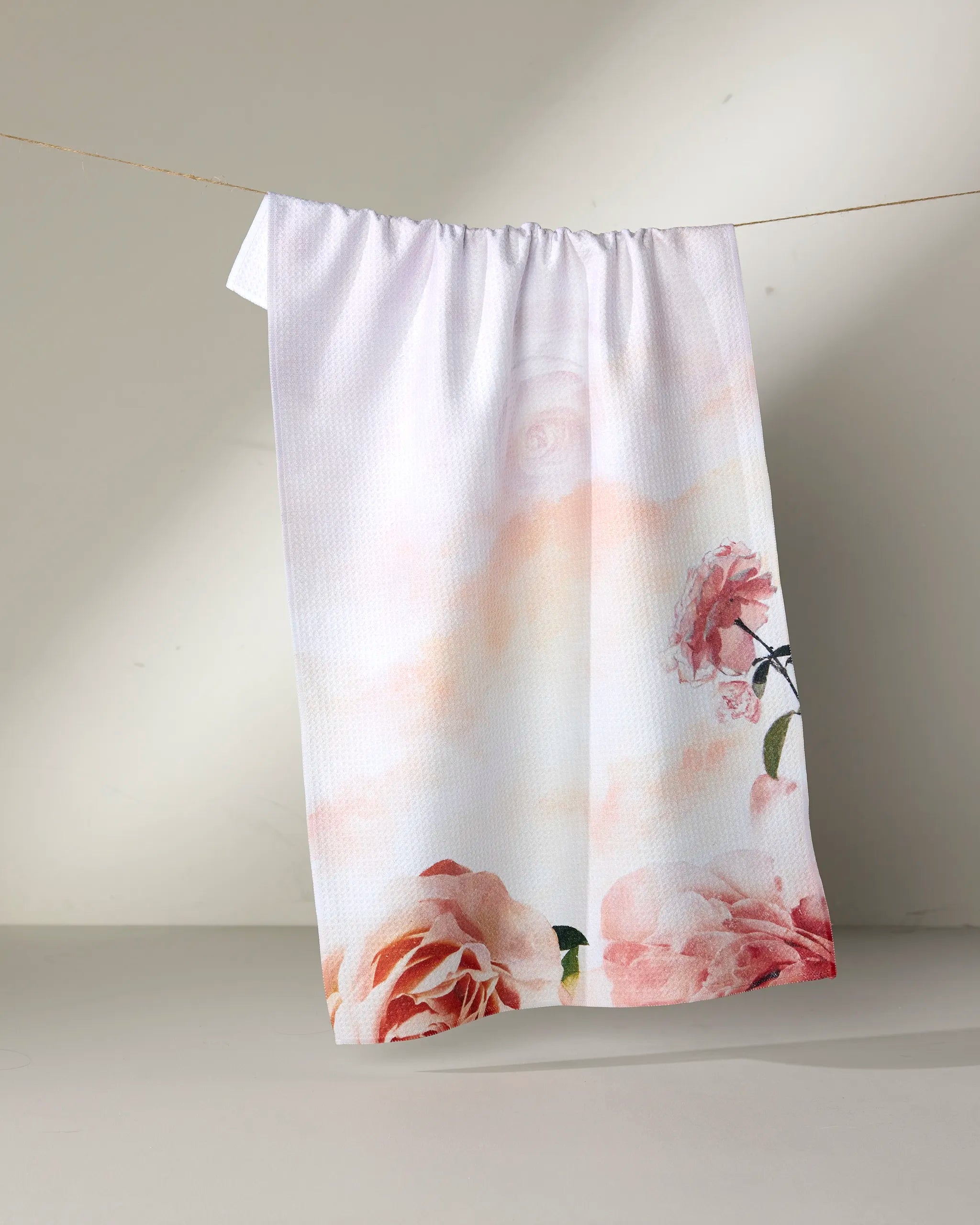 The Rhapsody in Bloom tea towel by Stefanie Bales features a delicate pink floral cluster at the bottom corner, hanging on a clothesline against a neutral background. Soft light casts gentle shadows on this ultra-absorbent kitchen accessory.