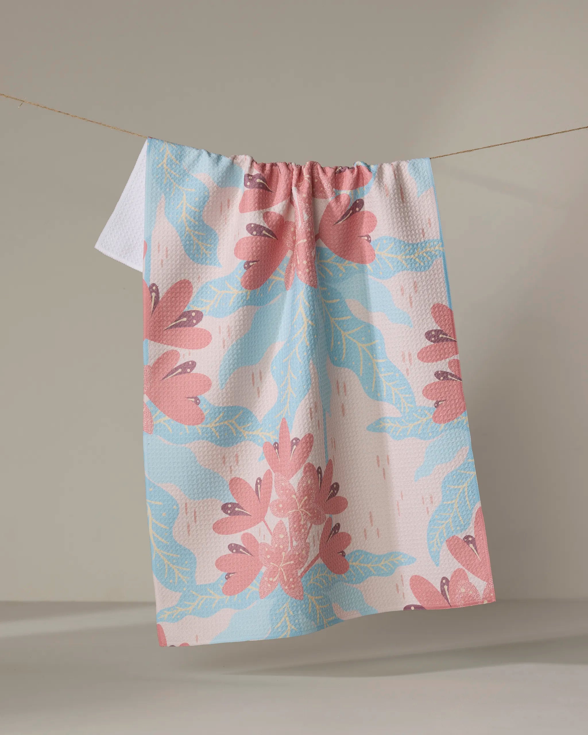 The Rhododendron kitchen tea towel by Julianne Haness, featuring a pink and blue floral design, hangs on a clothesline against a neutral background.