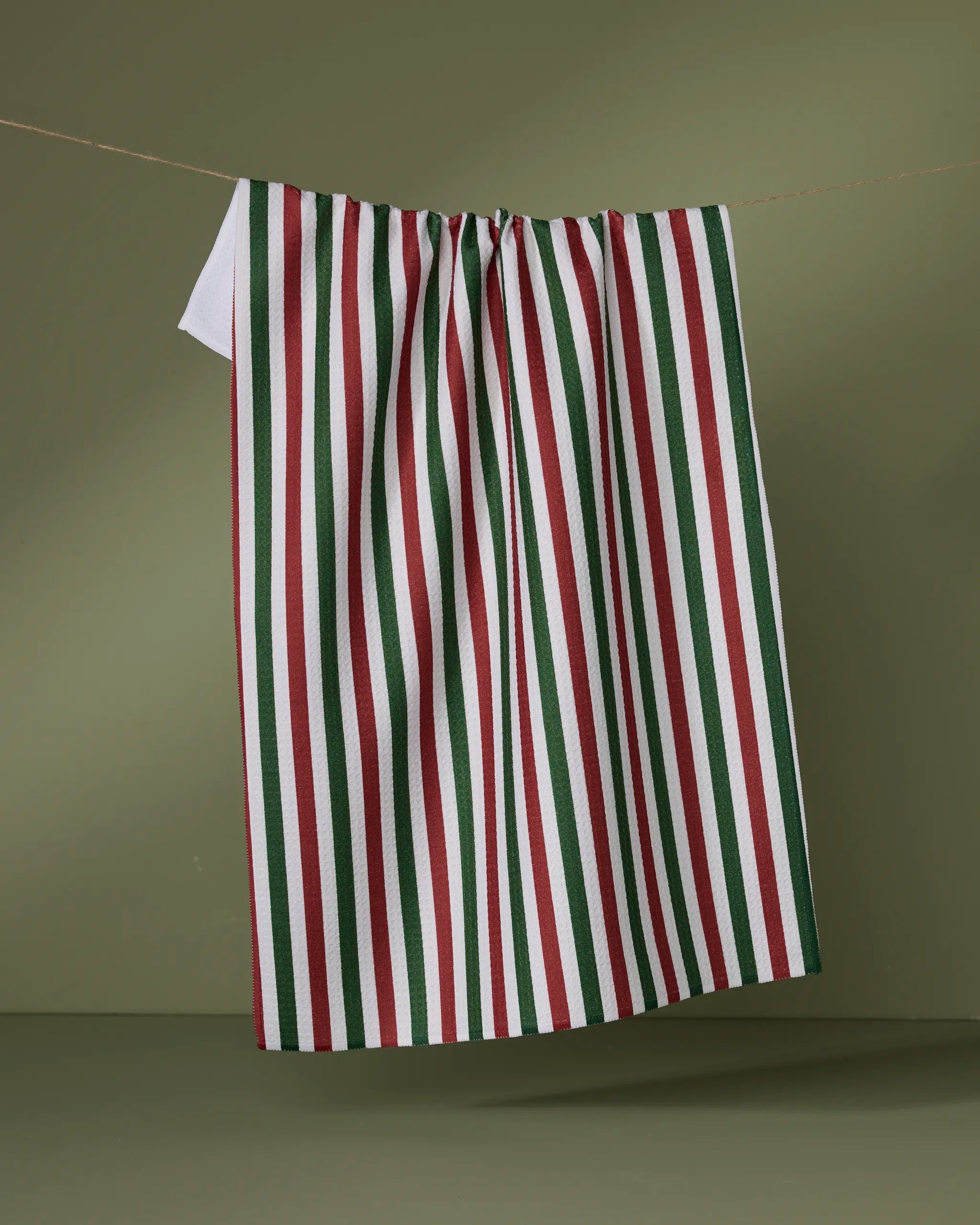 The Geometry Ribbon Green Red tea towel, featuring quick-drying fabric and vertical red, green, and white stripes, hangs on a clothesline against a simple olive-green background.