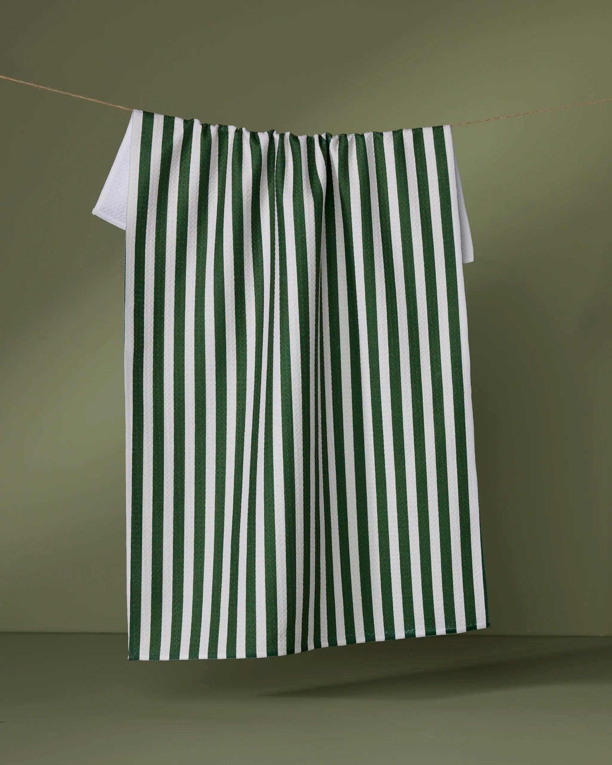 The Geometry Ribbon Green tea towel, made from recycled materials, features vertical green stripes on white and hangs on a string against an olive green background.