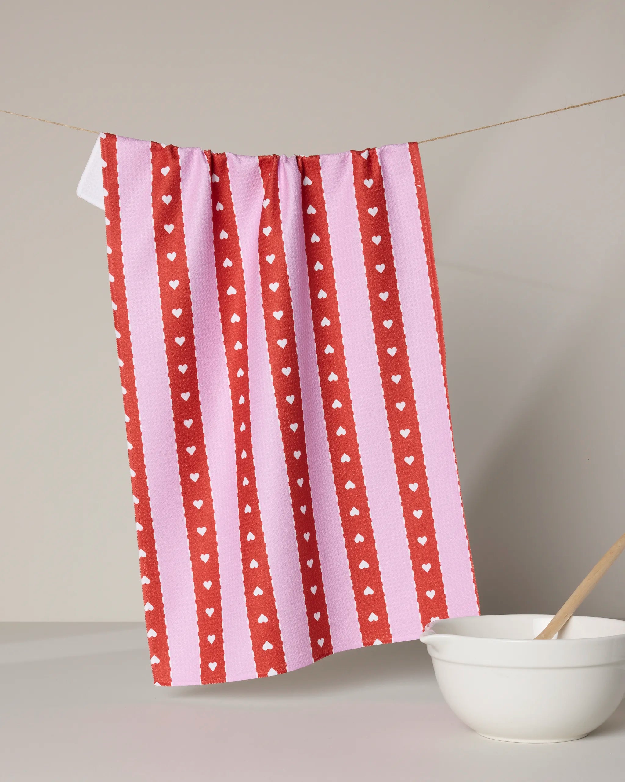 The Geometry Ribbon Hearts tea towel, made from recycled materials and featuring pink and red vertical stripes with white hearts, hangs on a clothesline above a white bowl with a wooden spoon on a light gray surface.