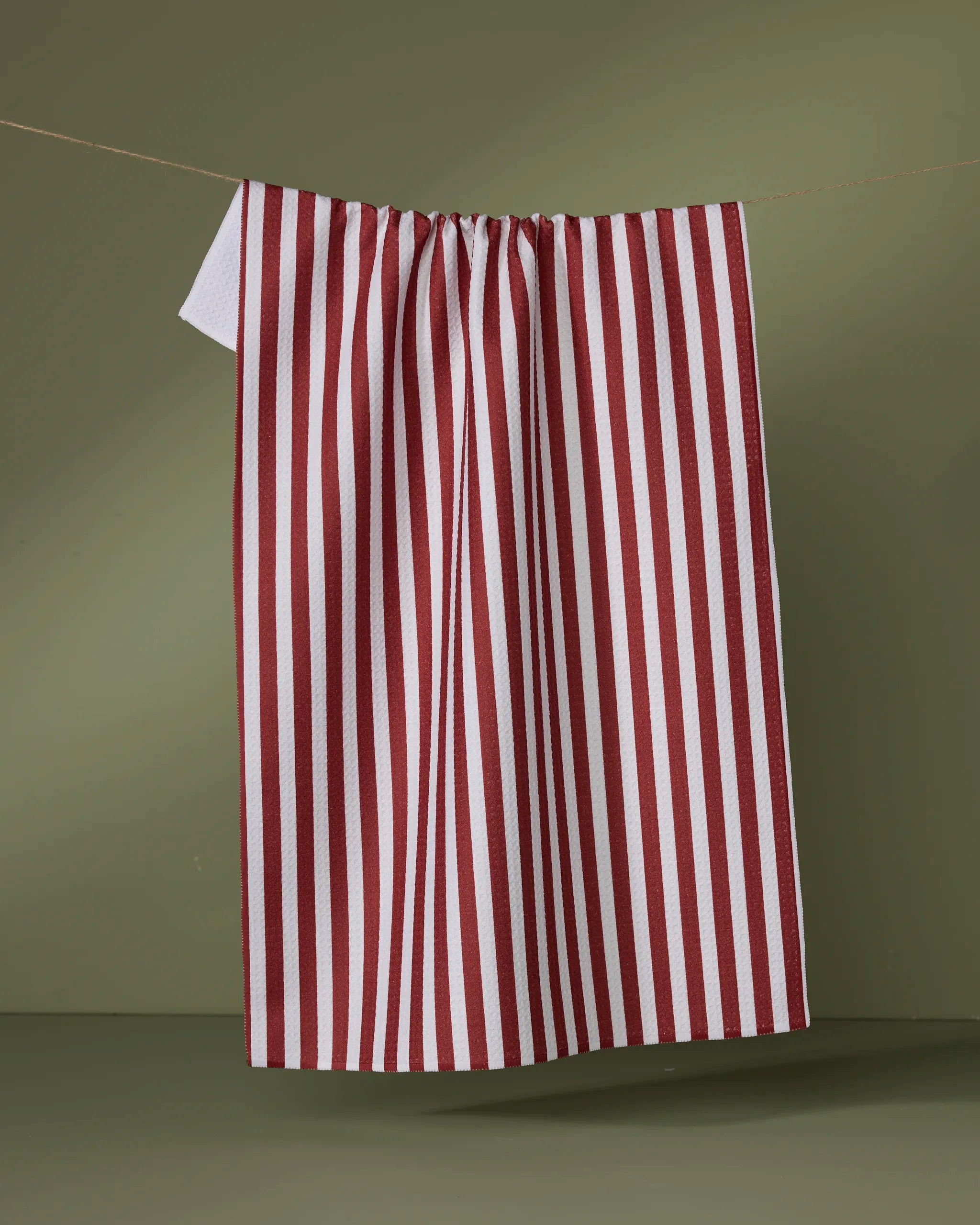 The Geometry Ribbon Red tea towel, featuring red and white vertical stripes and made from recycled materials, hangs on a string against a plain olive green background.