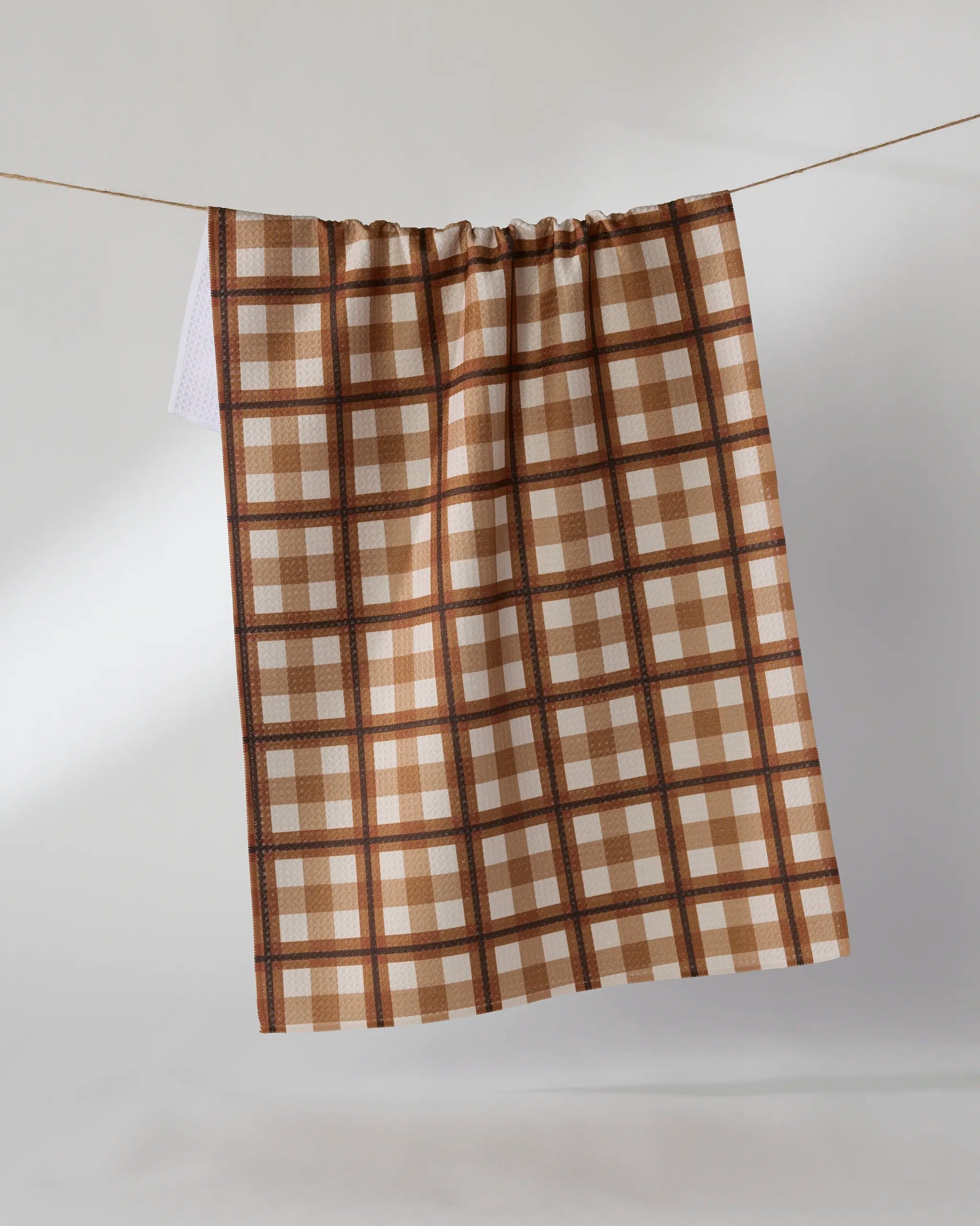 The Geometry "Rich Cocoa" tea towel, featuring a brown, beige, and white checkered pattern and made from recycled materials, hangs from a clothesline against a plain light background.