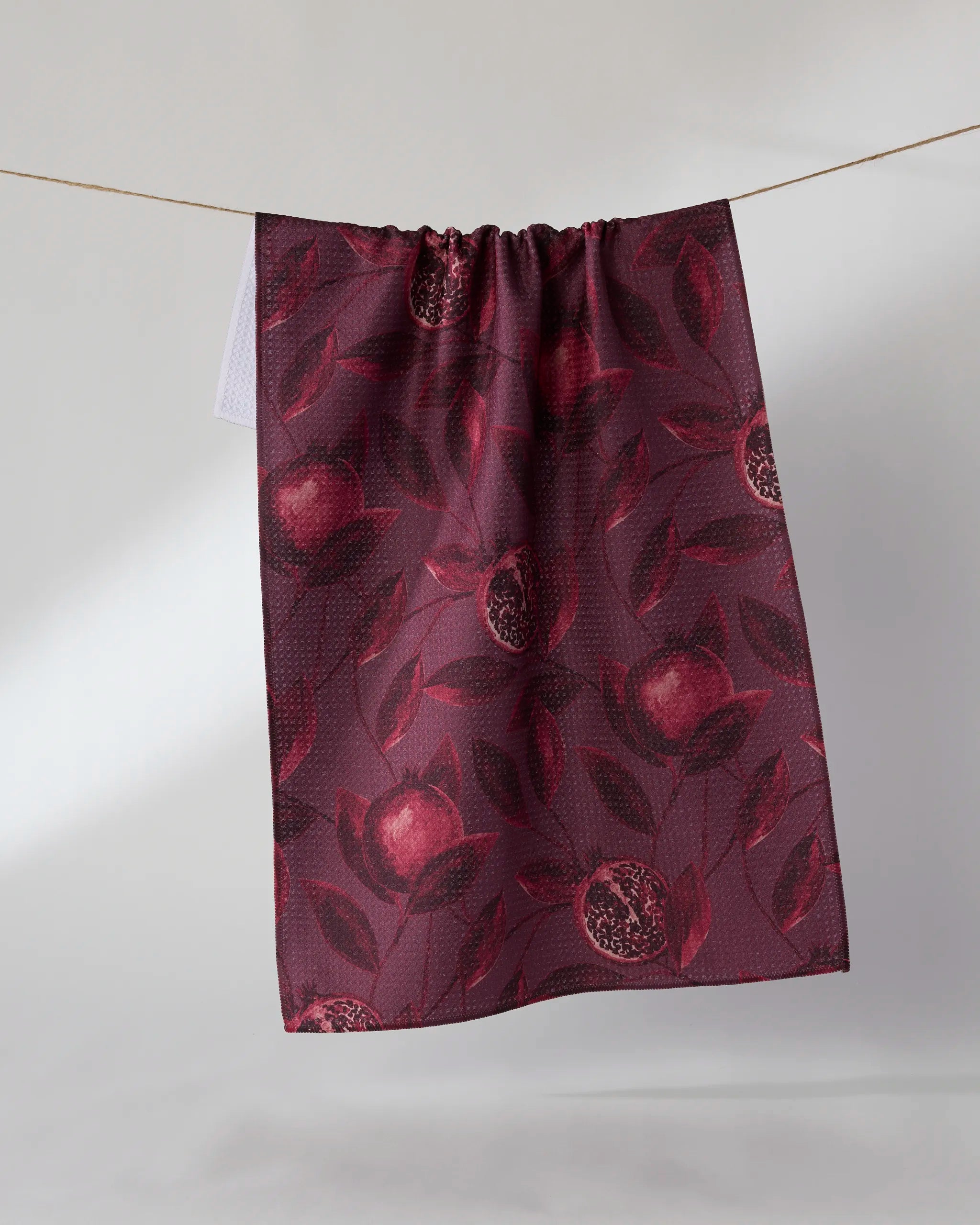 The Geometry "Rich Garnet" tea towel, featuring a pomegranate and leafy pattern in deep red, dries quickly and hangs on a clothesline against a simple light background.