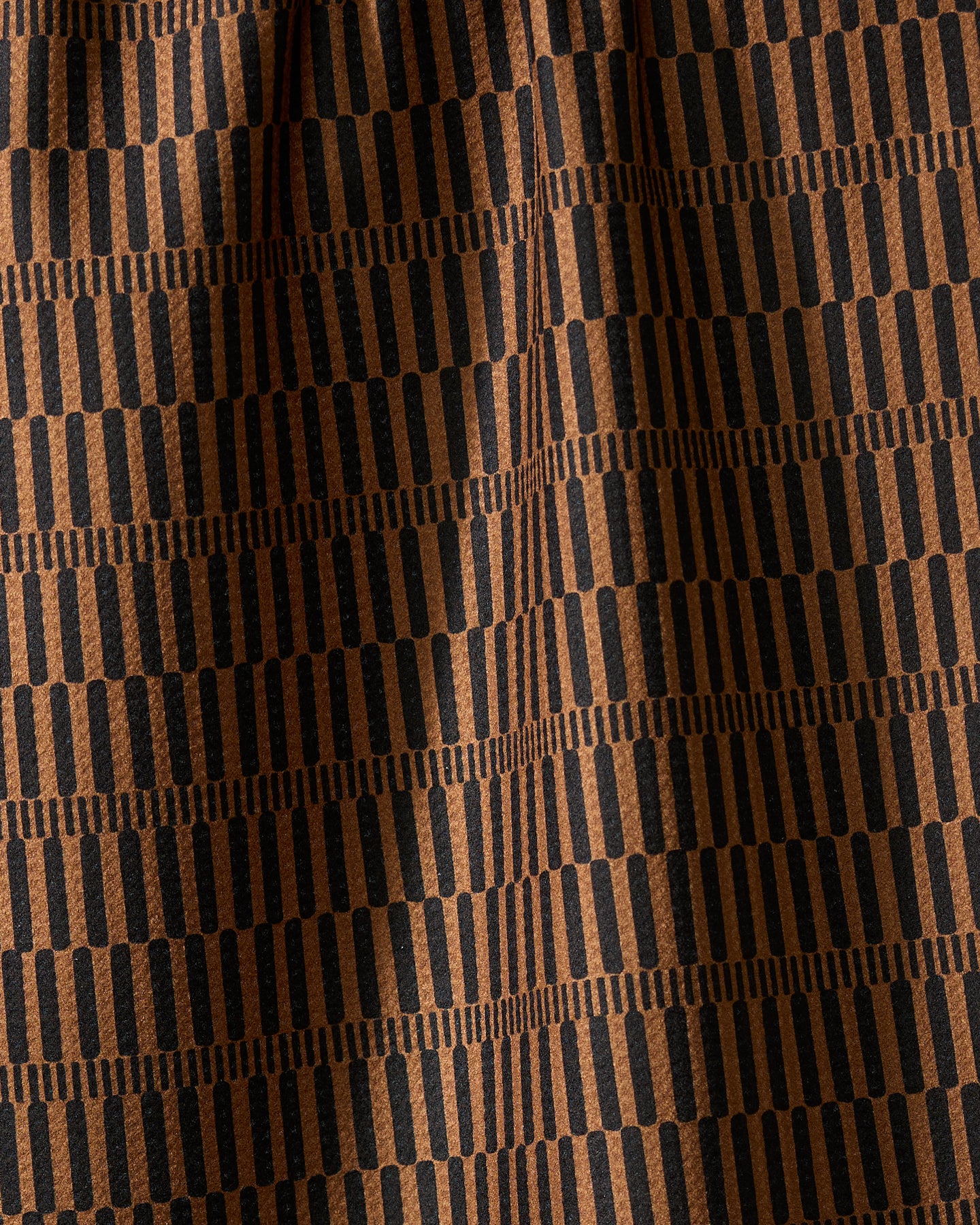 Ridge by Geometry is a recycled kitchen towel featuring a repeating pattern of vertical black lines on a brown background, arranged in horizontal rows. Its slightly wrinkled texture creates soft folds and shadows.