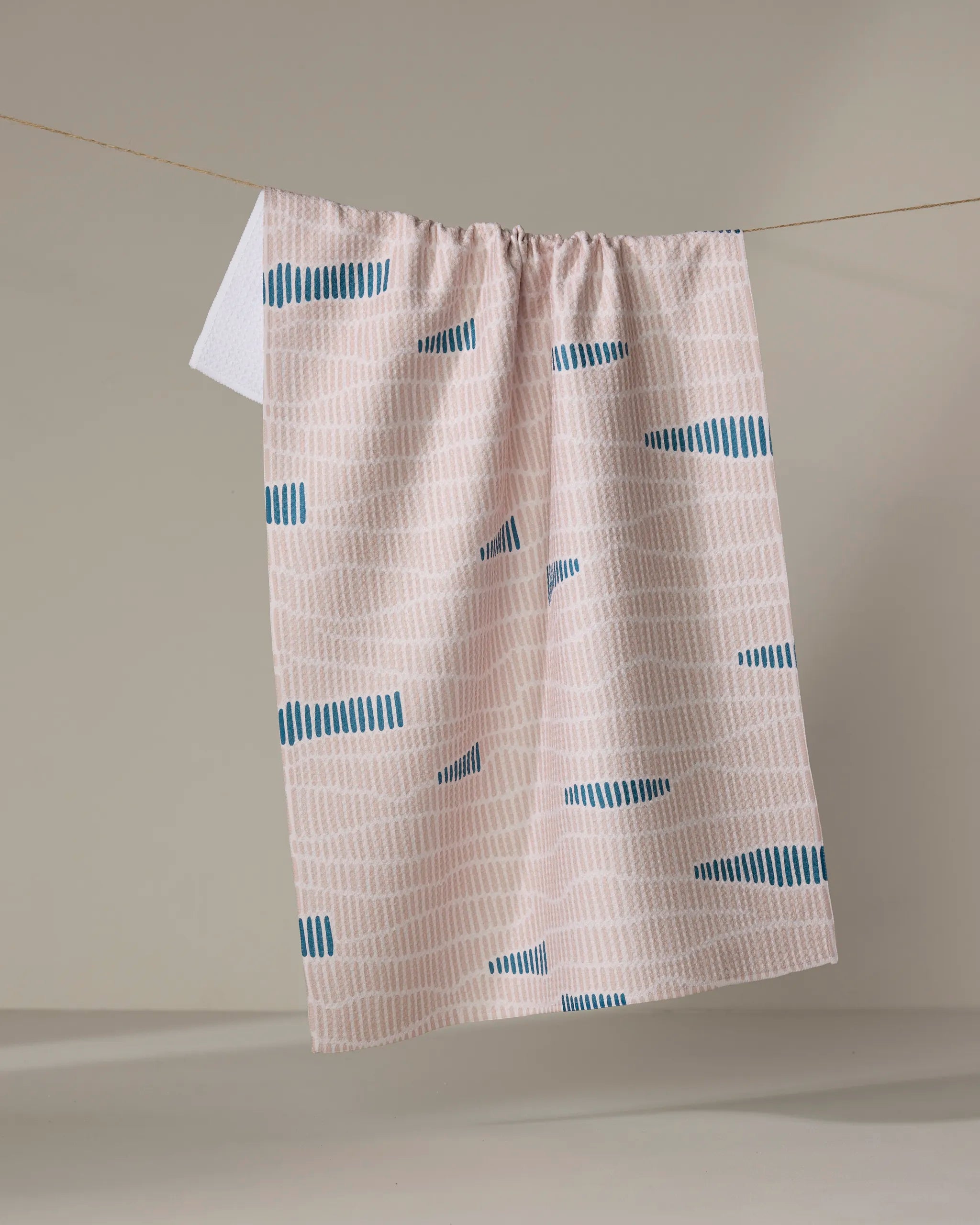 The Twig + Spark Ridgeline kitchen tea towel, made from recycled materials, features light pink fabric with vertical beige lines and blue dashes, displayed hanging on a clothesline against a neutral background.